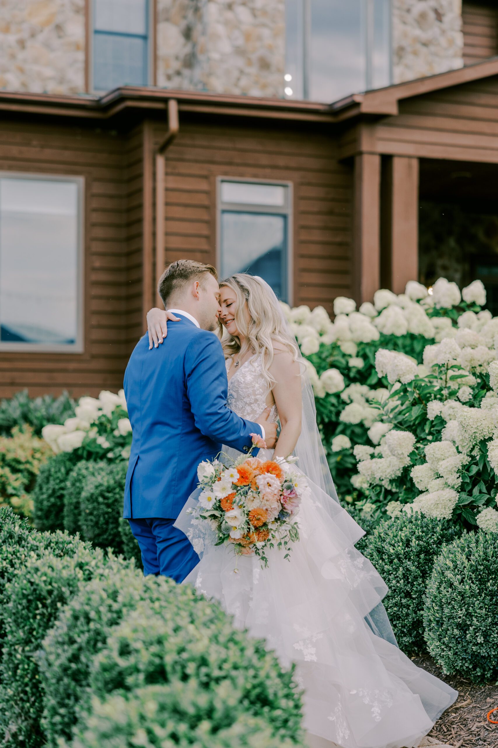 Bride and groom standing close with their noses touching, bride holding bouquet of orange, white, and pink flowers