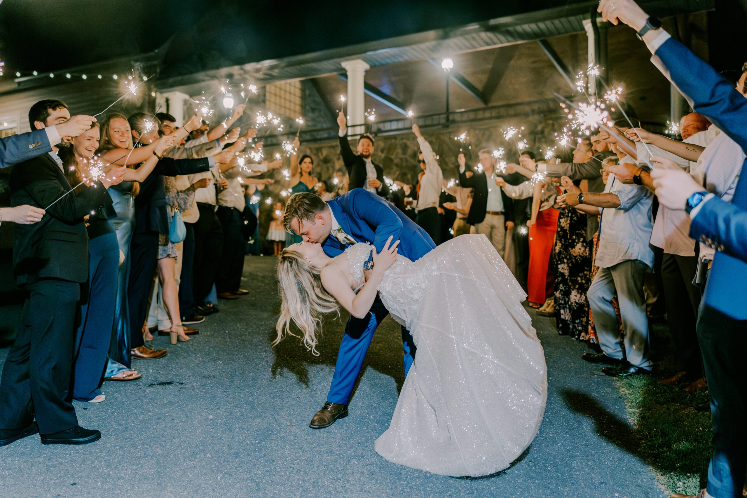 Groom dips bride in a kiss as they exit their wedding, guests surrounding them holding up sparklers and cheering them on