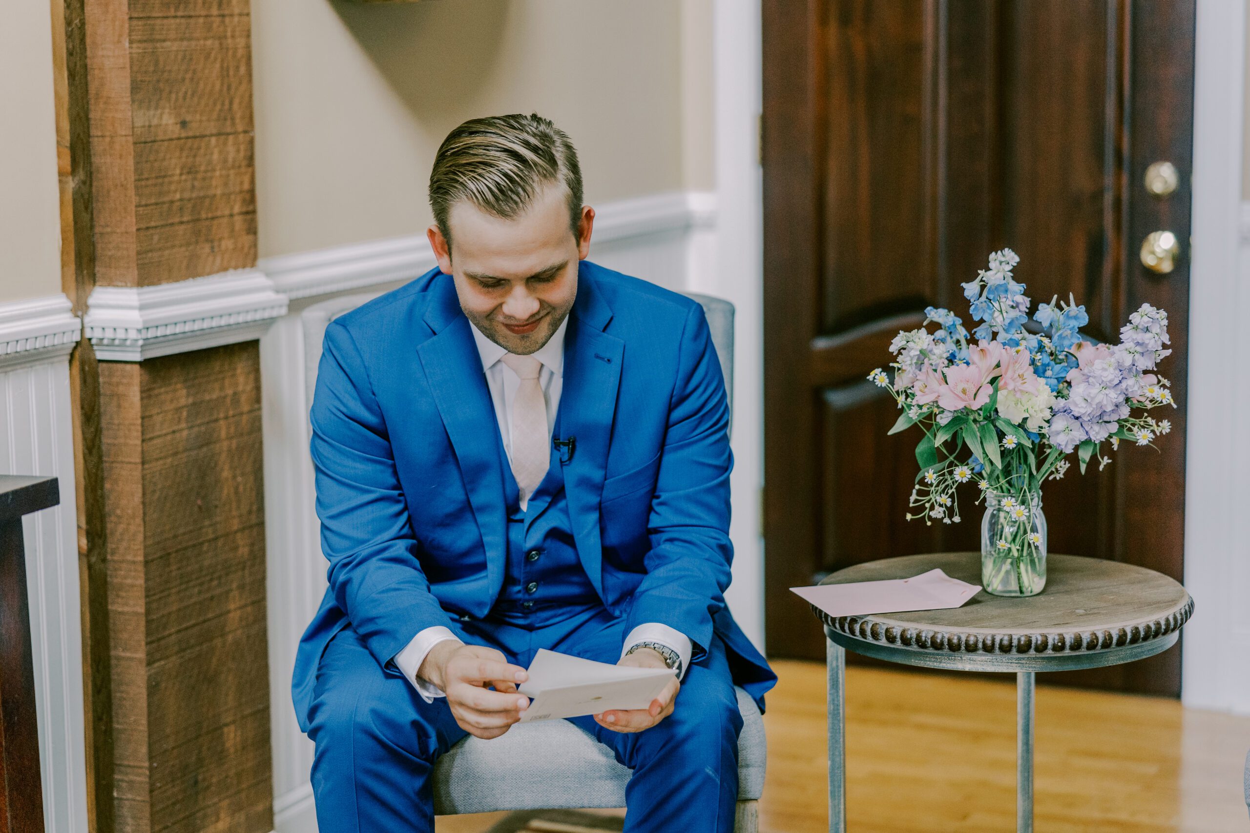 Groom sitting on a chair in a blue suit reading a card while smiling