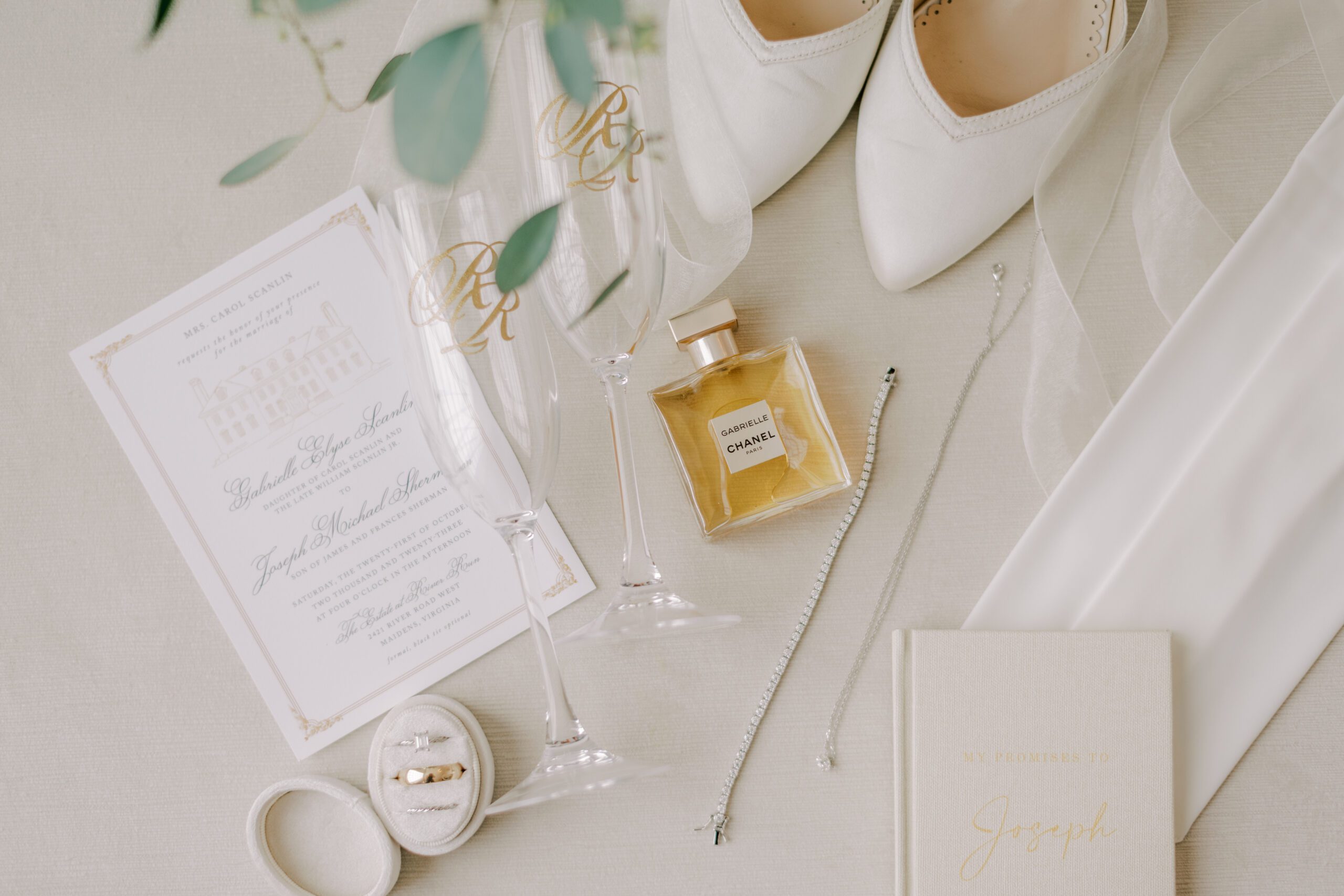 Detail photo of bride's perfume, shoes, jewelry, vow book, champagne glasses, and the wedding invitation