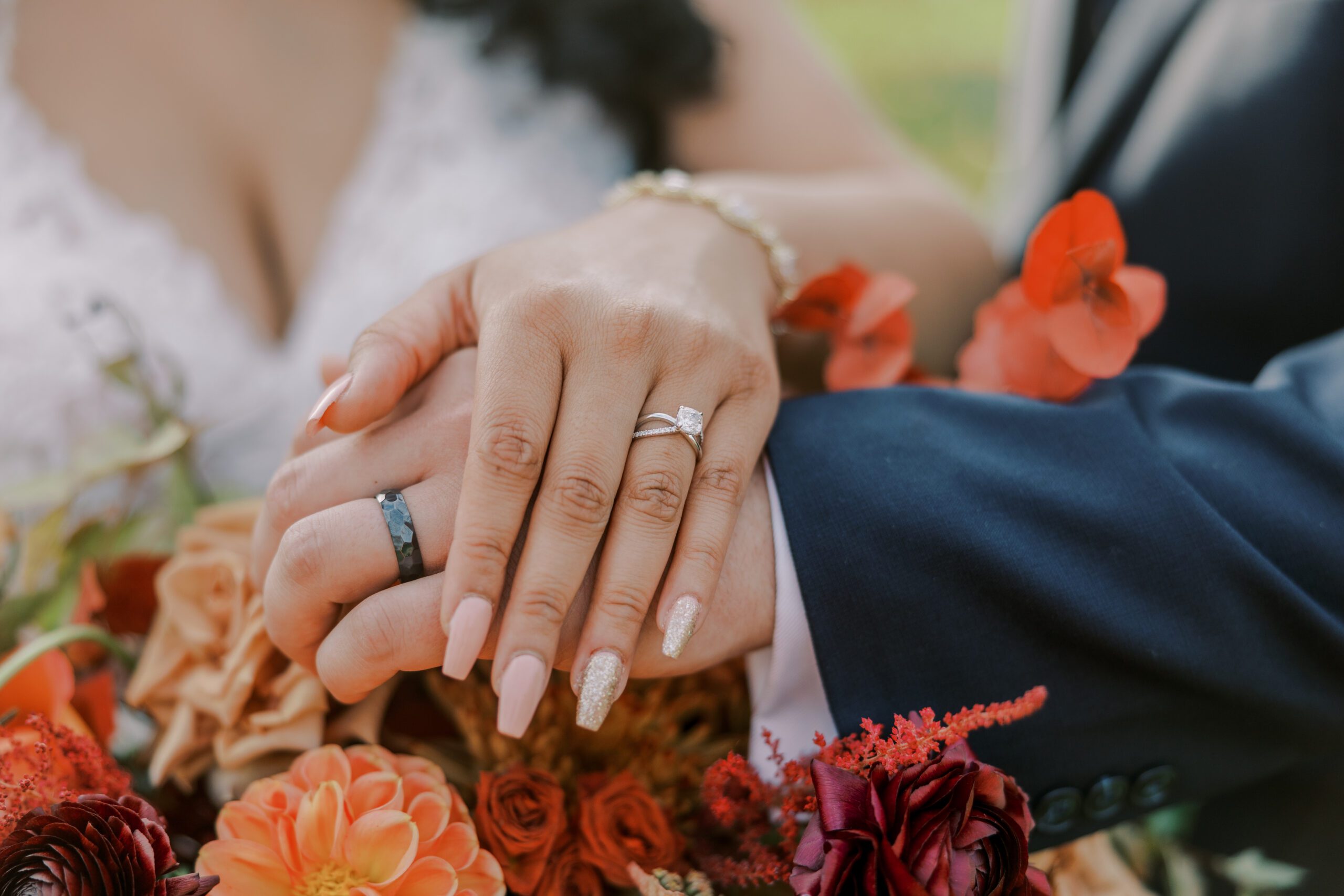 Close up photo of bride and groom's hands, bride has her double banded engagement ring on, and groom has a faceted black band ring, bride's nails are a pink tinted cream color and her ring finger nail is sparkly
