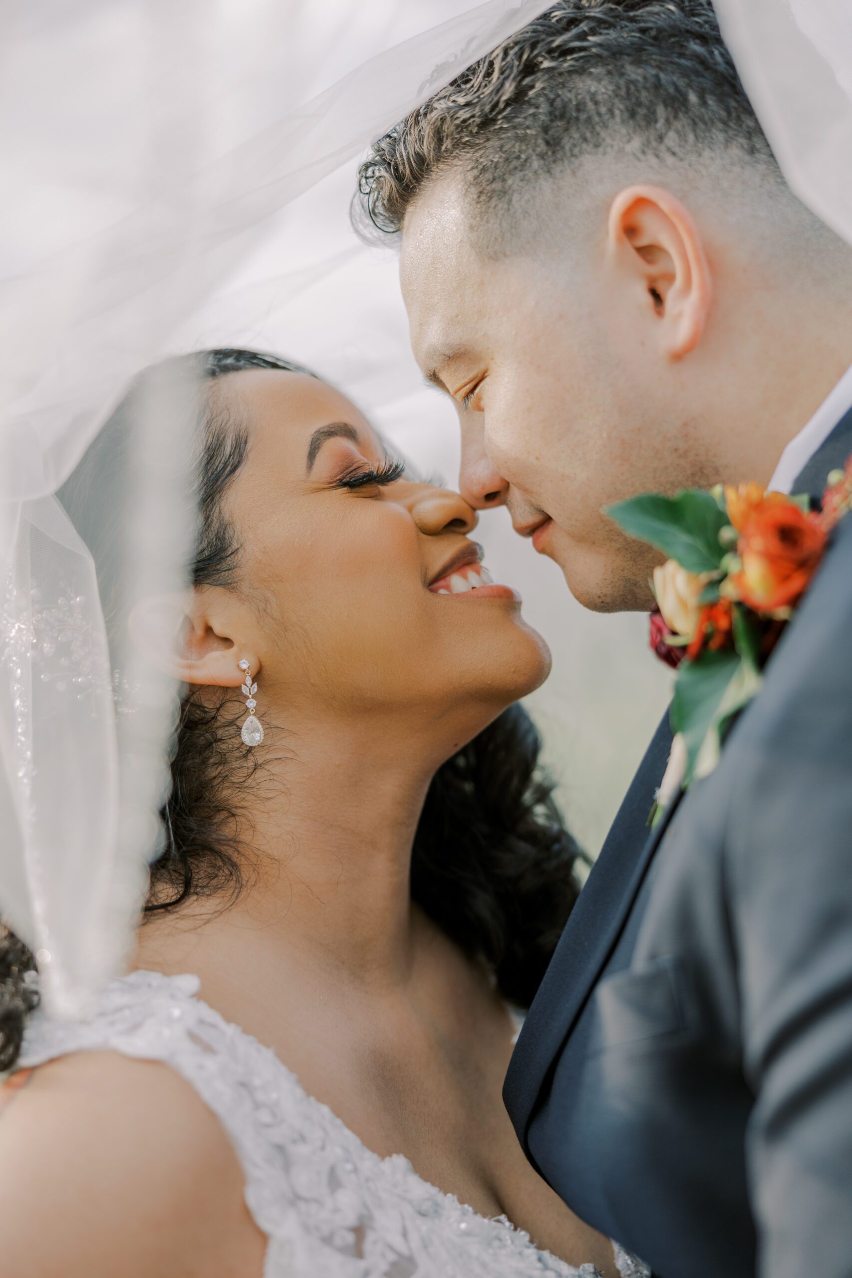 Close up image of bride and groom touching noses and smiling, bride's veil over both of their heads