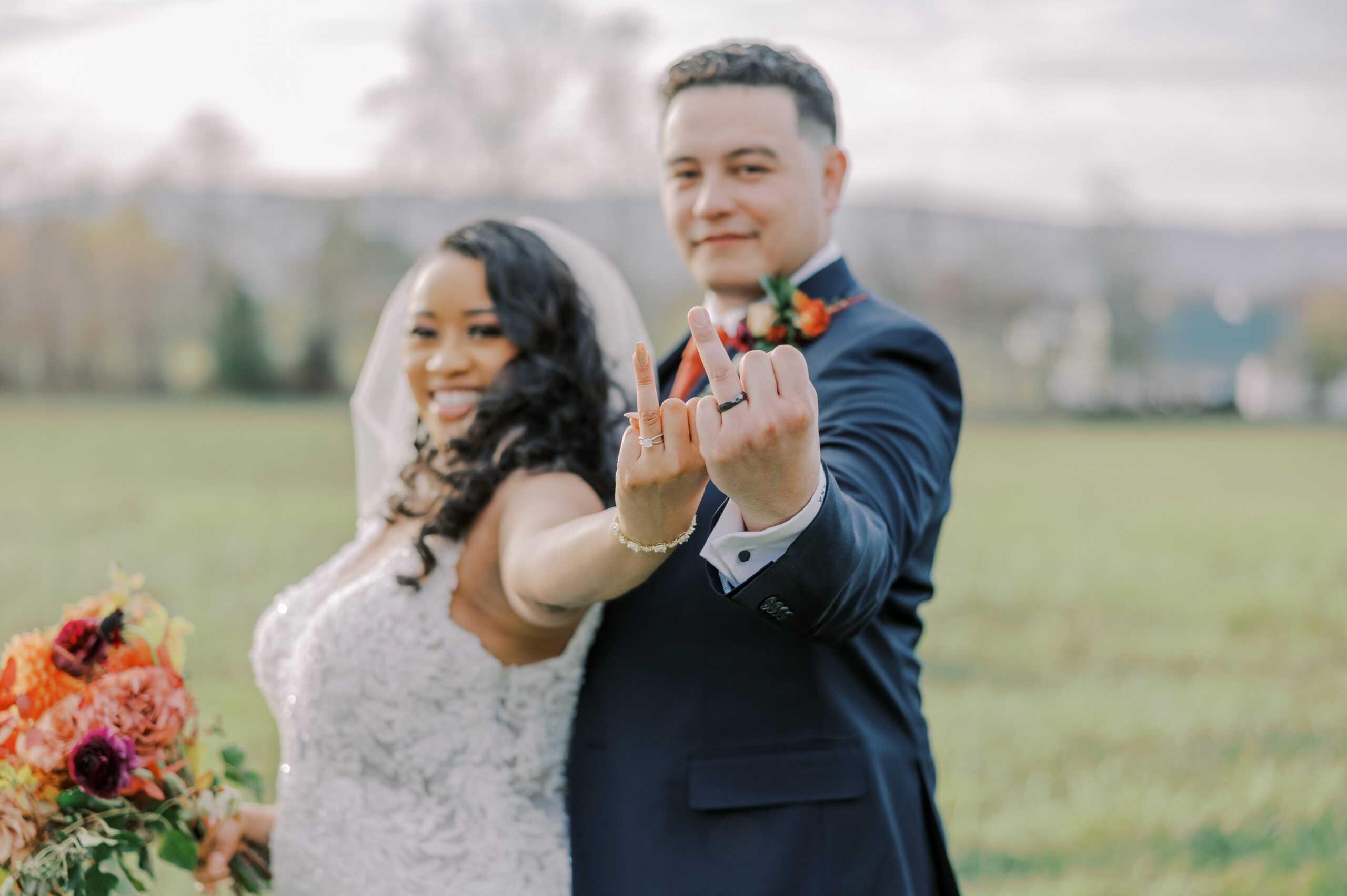 Photo of bride and groom holding up their ring fingers showing off their rings, couple is slightly out of focus, hands in focus