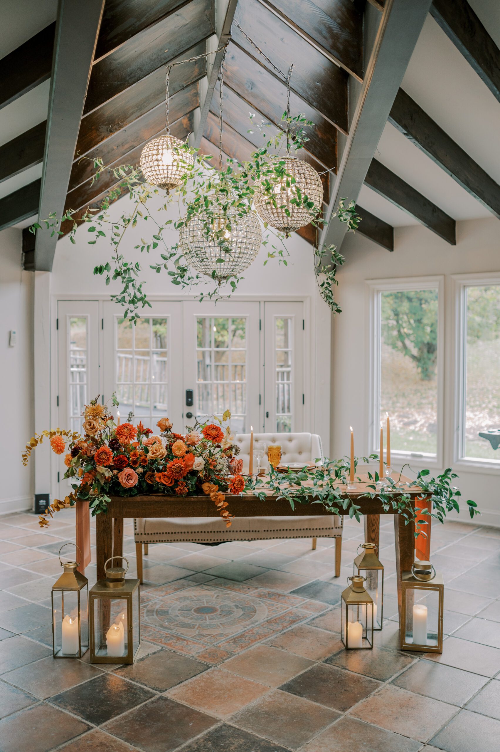 Interior photo of the manor at airmont of bride and groom's sweetheart table, covered in flowers and candles, with greenery and 3 large rounded hanging lights above