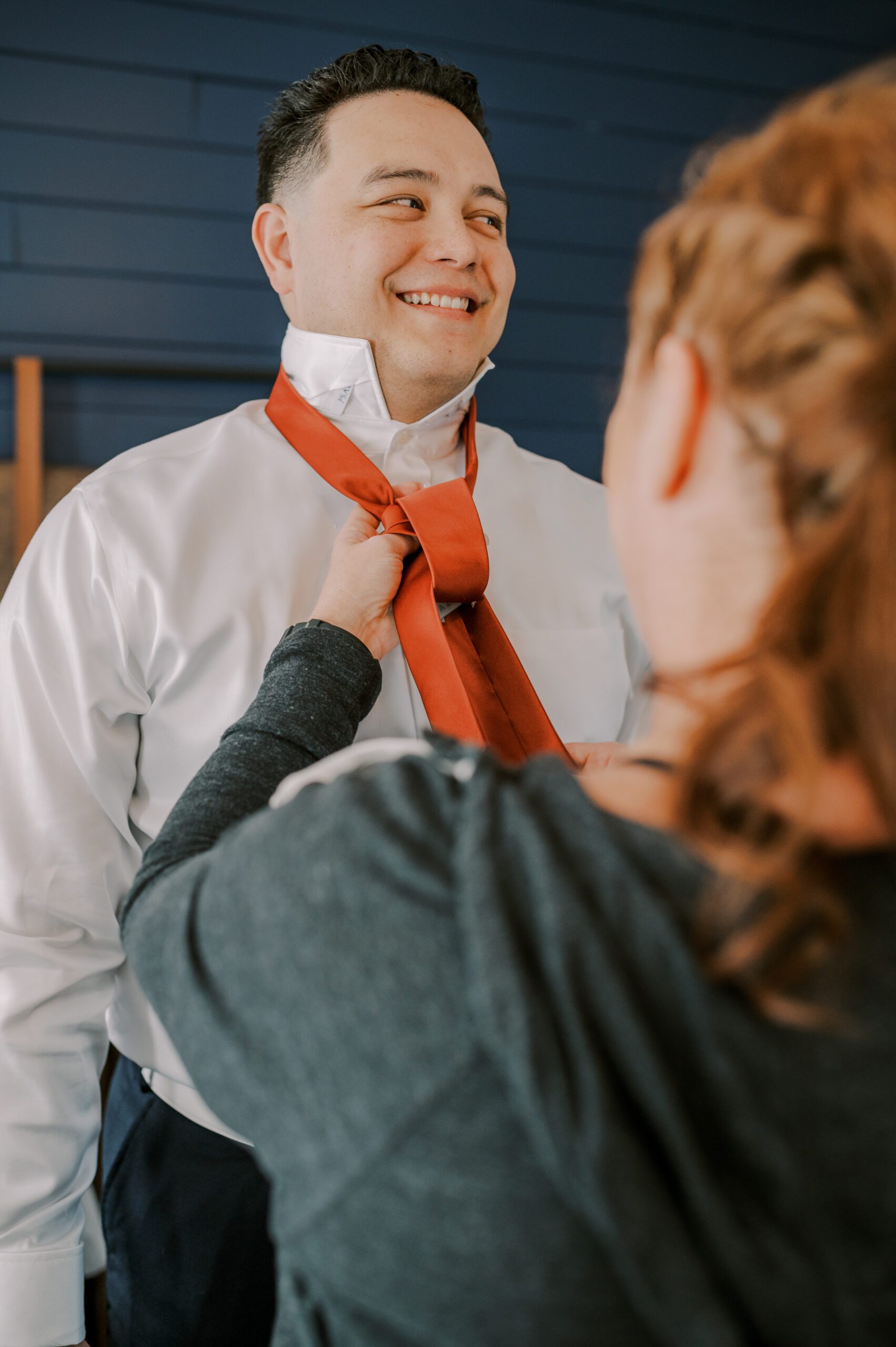 Groom smiling off to side while a woman ties his orange tie for him