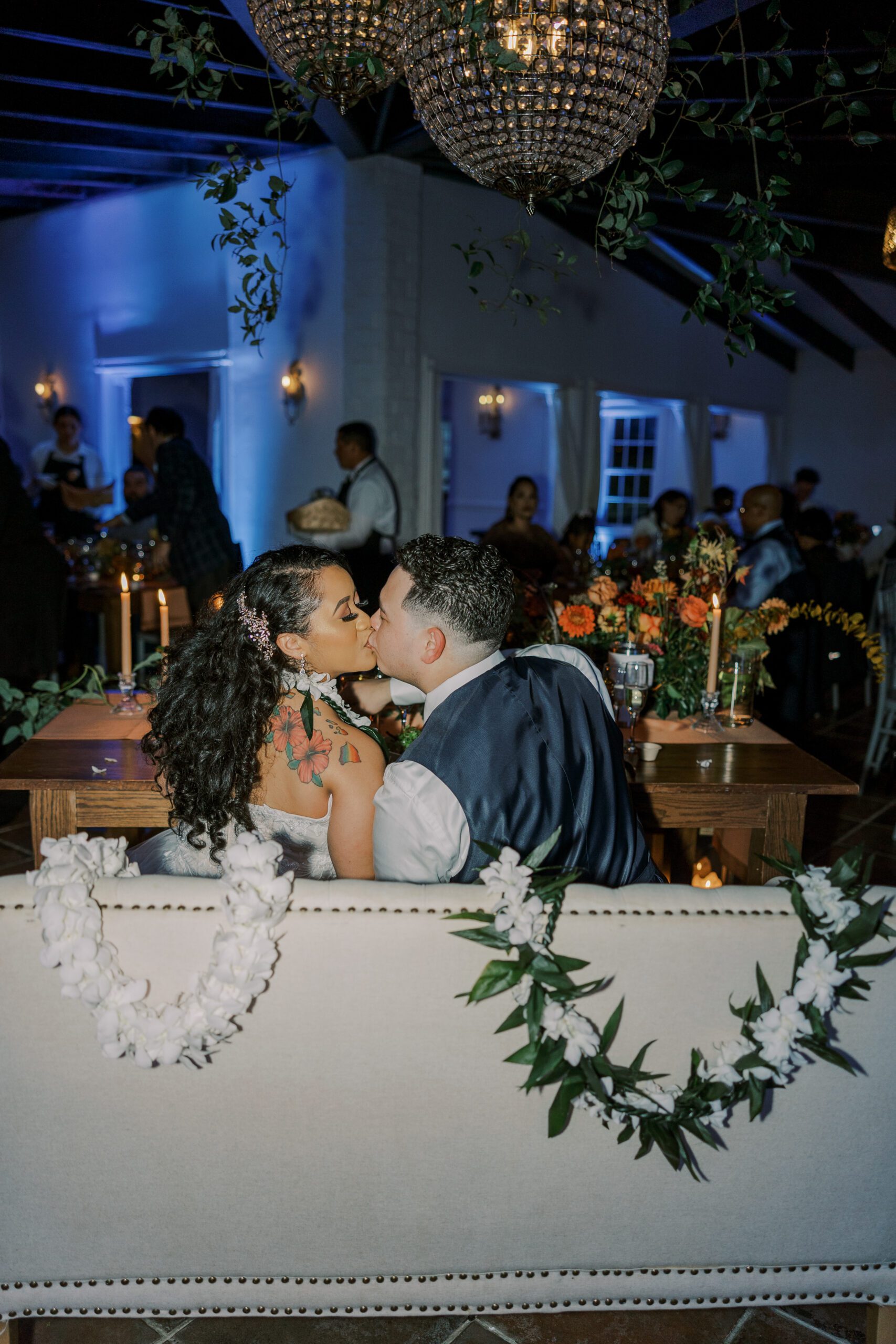 Couple kissing while they sit at their reception table, photo taken from behind with all their guests pictured in background