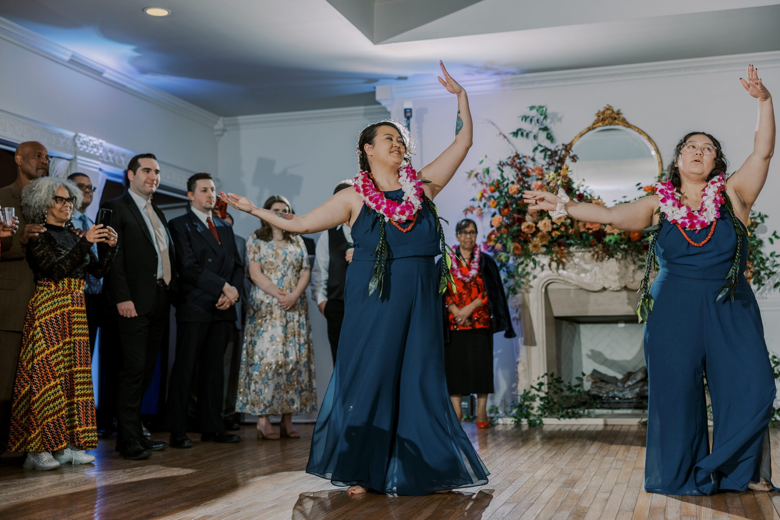 Two women in blue jumpsuits dancing in unison with their arms up, wearing leis