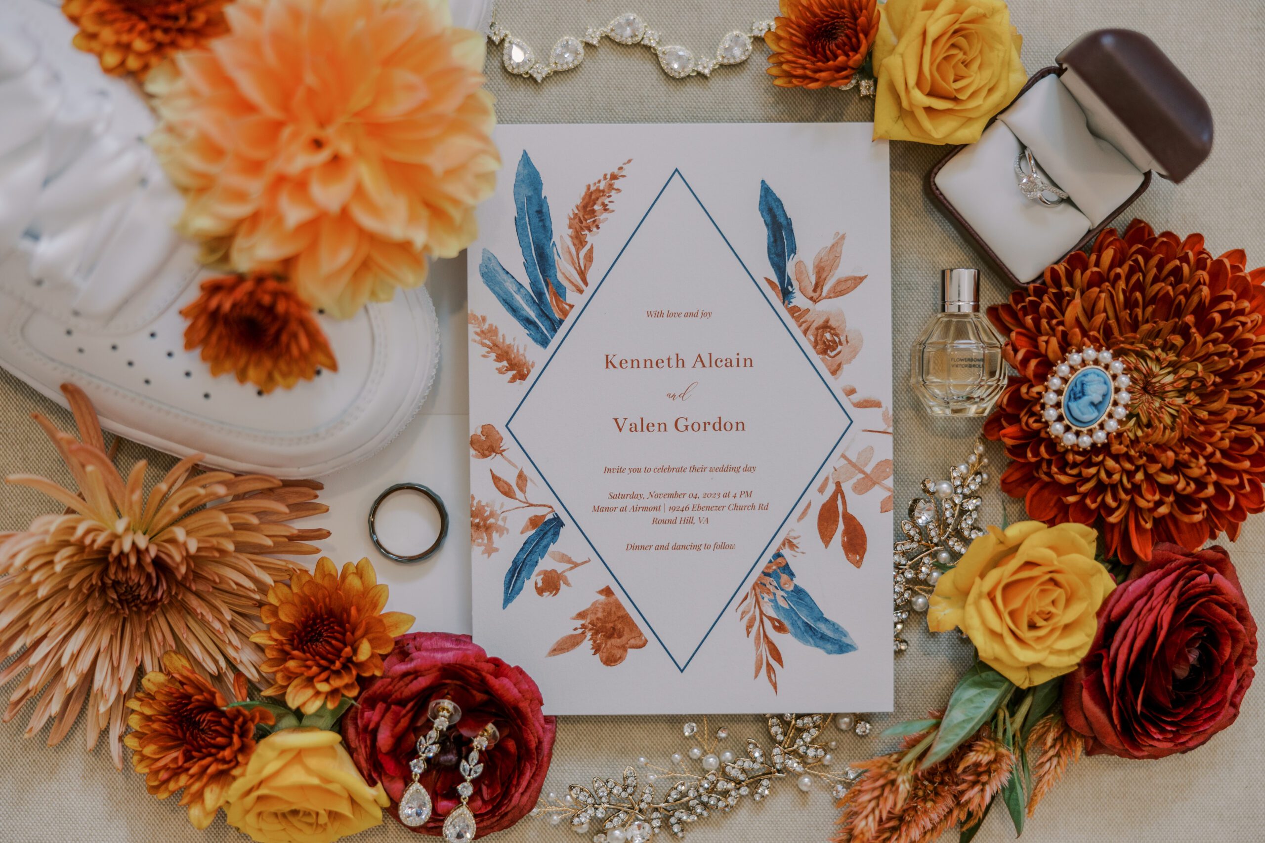 Detailed photo of wedding invitation on white paper with blue diamond shape, details written inside with orange and blue flowers and feathers, other florals, rings, and jewelry surround the invitation