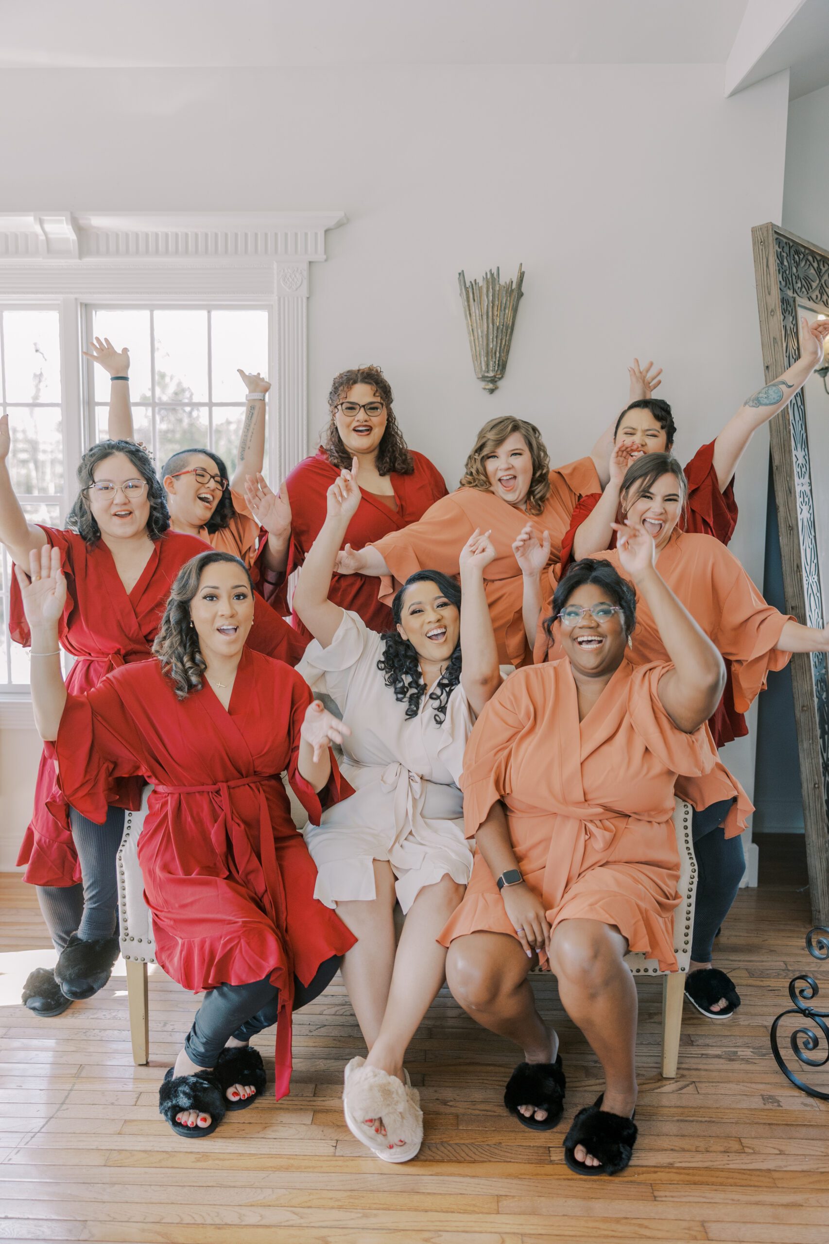 Group photo of bride wearing a white robe, and her bridesmaids wearing red and orange robes, all cheering with their hands up in the air at the manor at airmont fall wedding