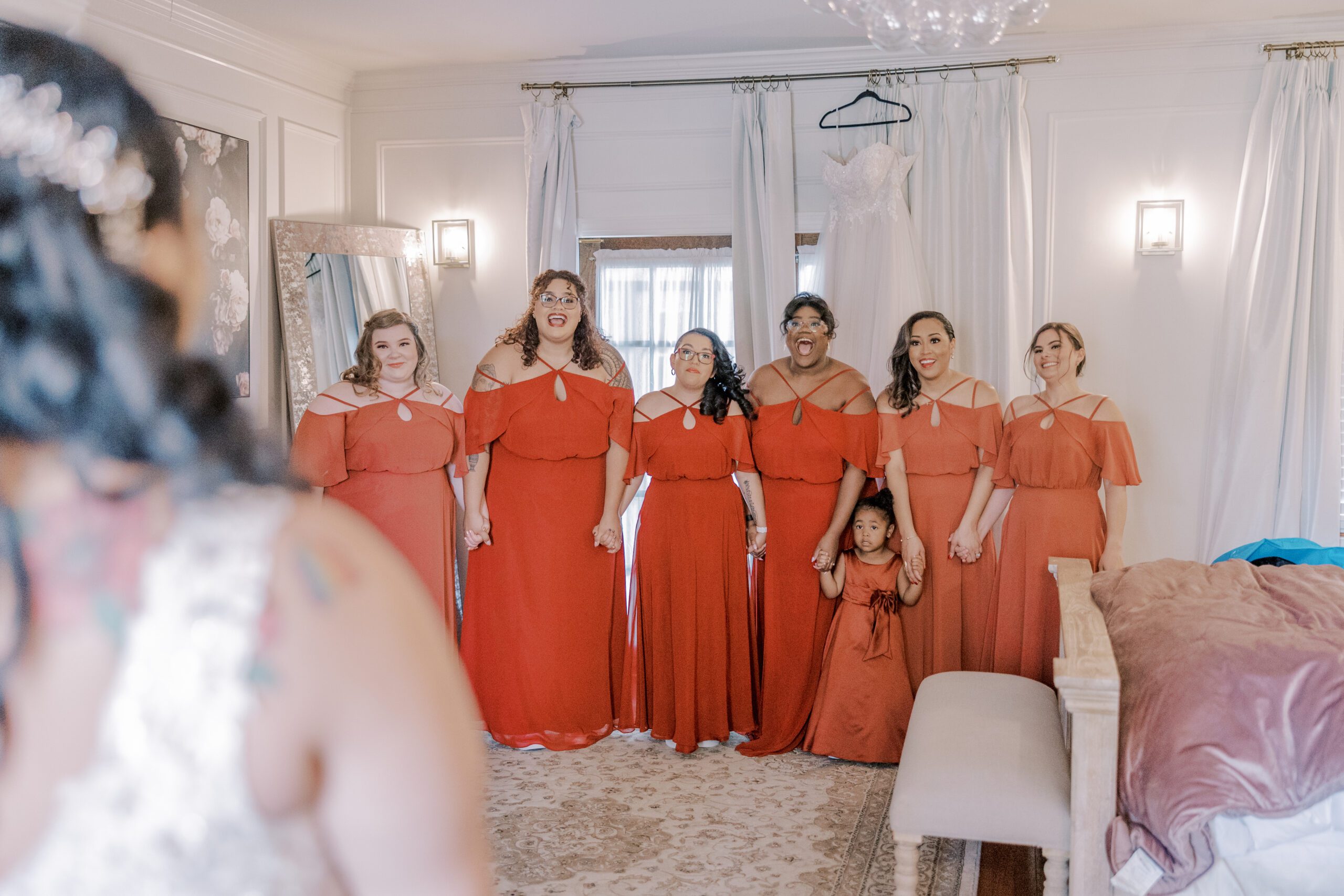 Bridesmaids in their orange dresses, along side a young girl also wearing an orange dress, all with excited expressions on their faces looking at the bride in her wedding gown