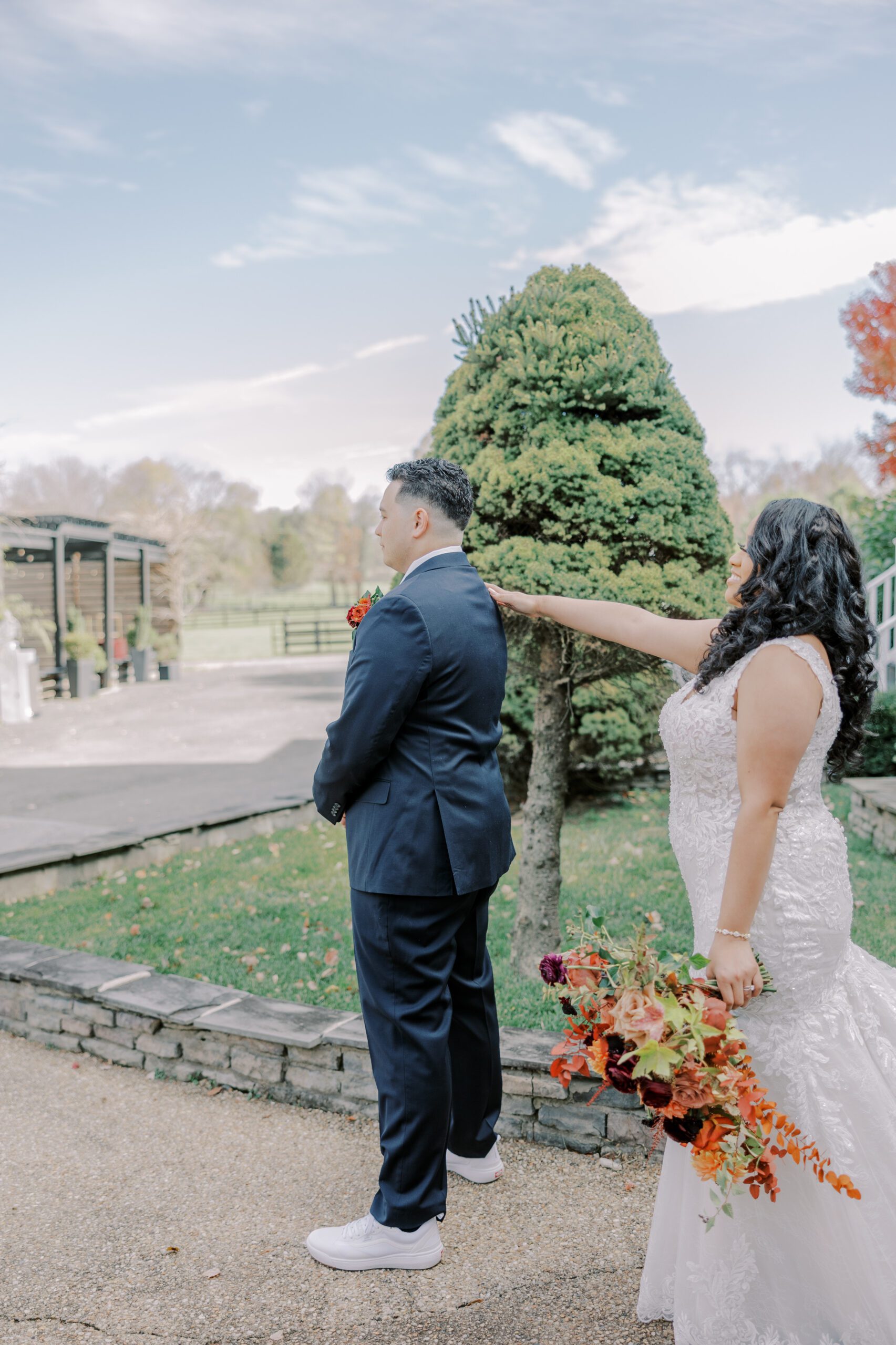 Bride outside tapping groom on shoulder while she holds her bouquet of orange, burgundy and red colored flowers