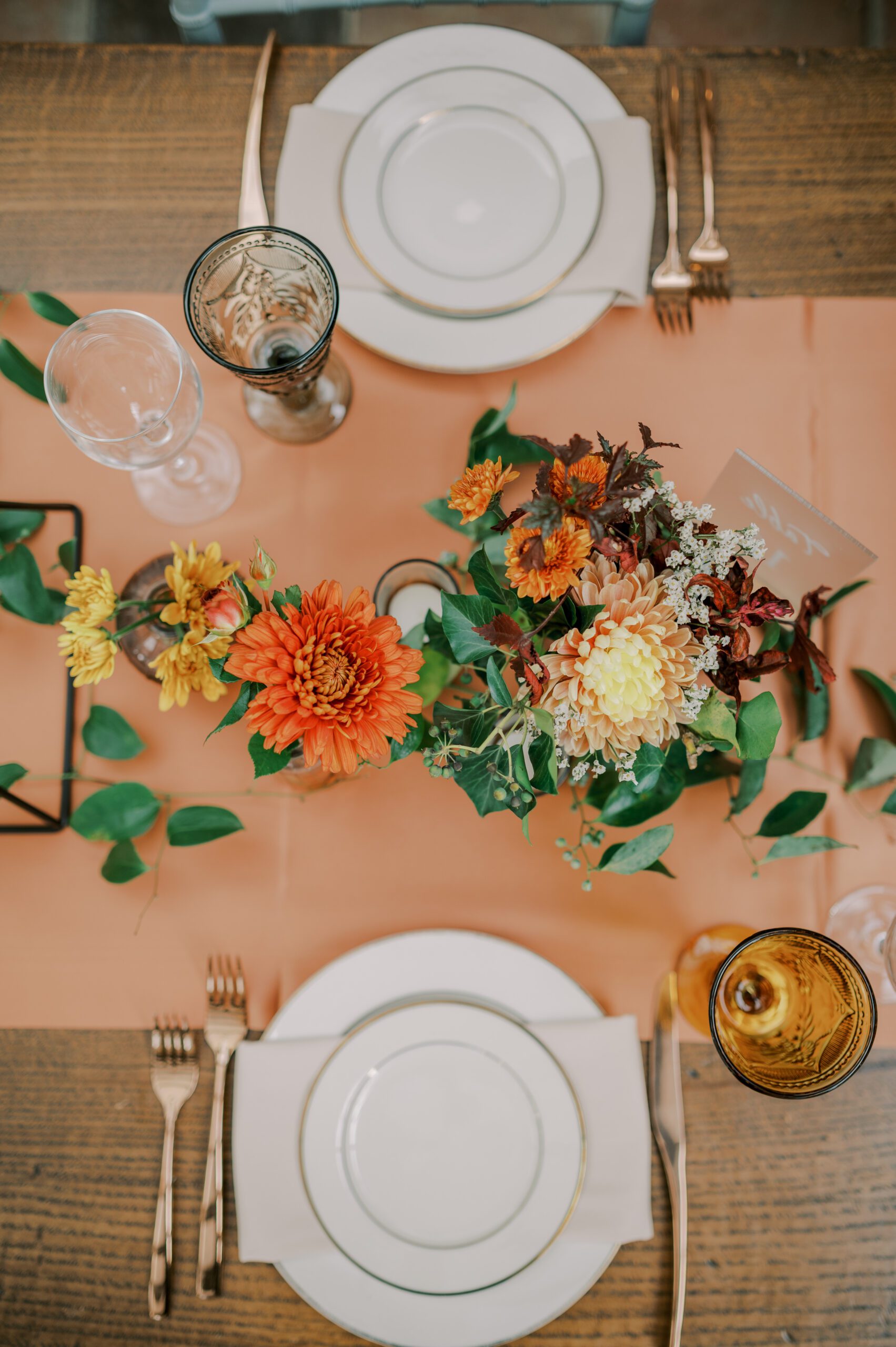 Bird's eye vide photo taken of table settings, white plates with gold rims, gold silverware, and orange, yellow and burgundy flowers in center