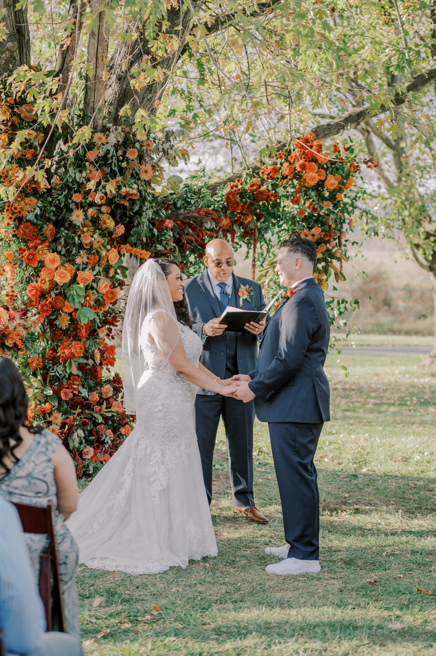 Bride and groom holding hands smiling at one another, tree covered in tons of red and orange flowers behind them