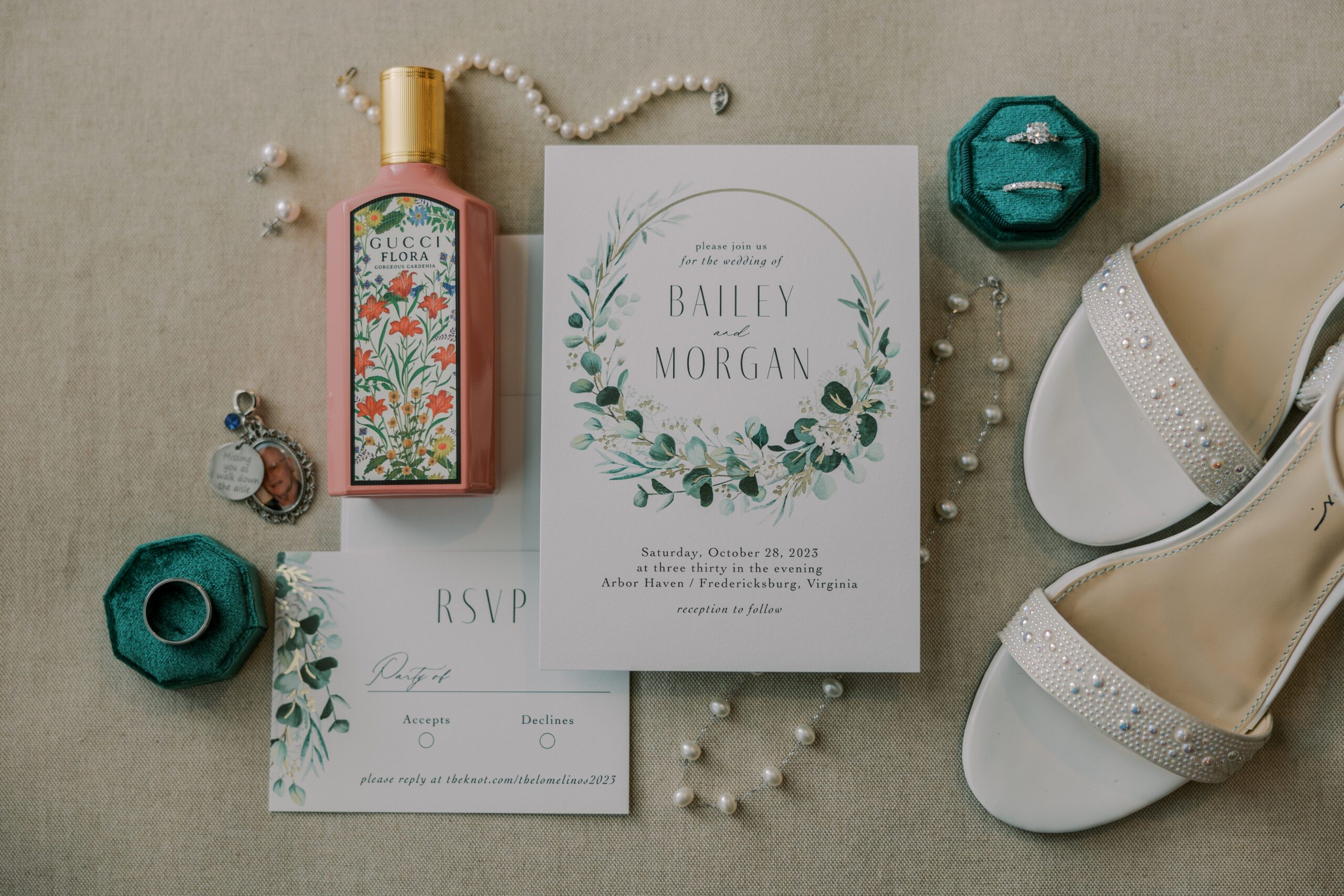 Detail photo of white wedding invitation with a gold circle and greenery on it, rsvp card, rings, bride's jewelry, shoes, bouquet charm, and perfume also pictured