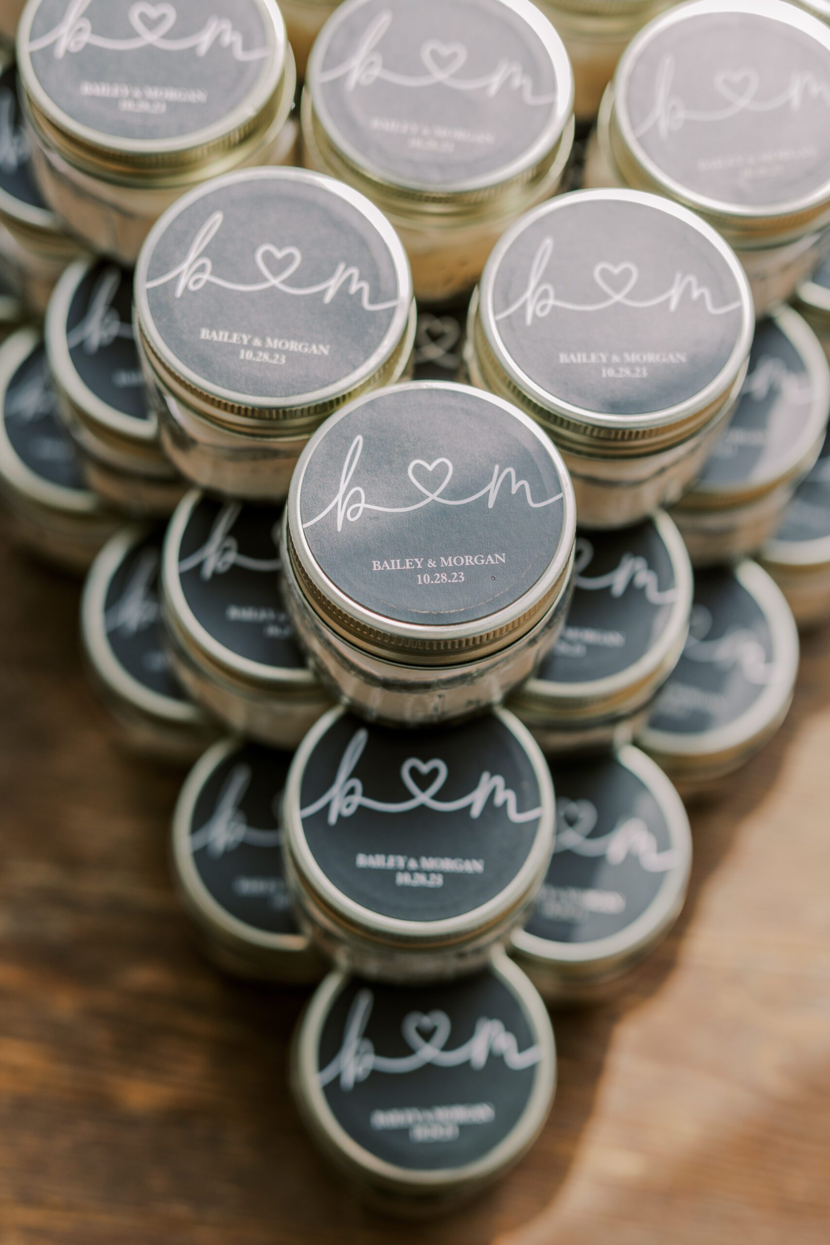 Large stack of little jars that have a black sticker on top that have a decorative b and m on them, as well as couple's names and their wedding date
