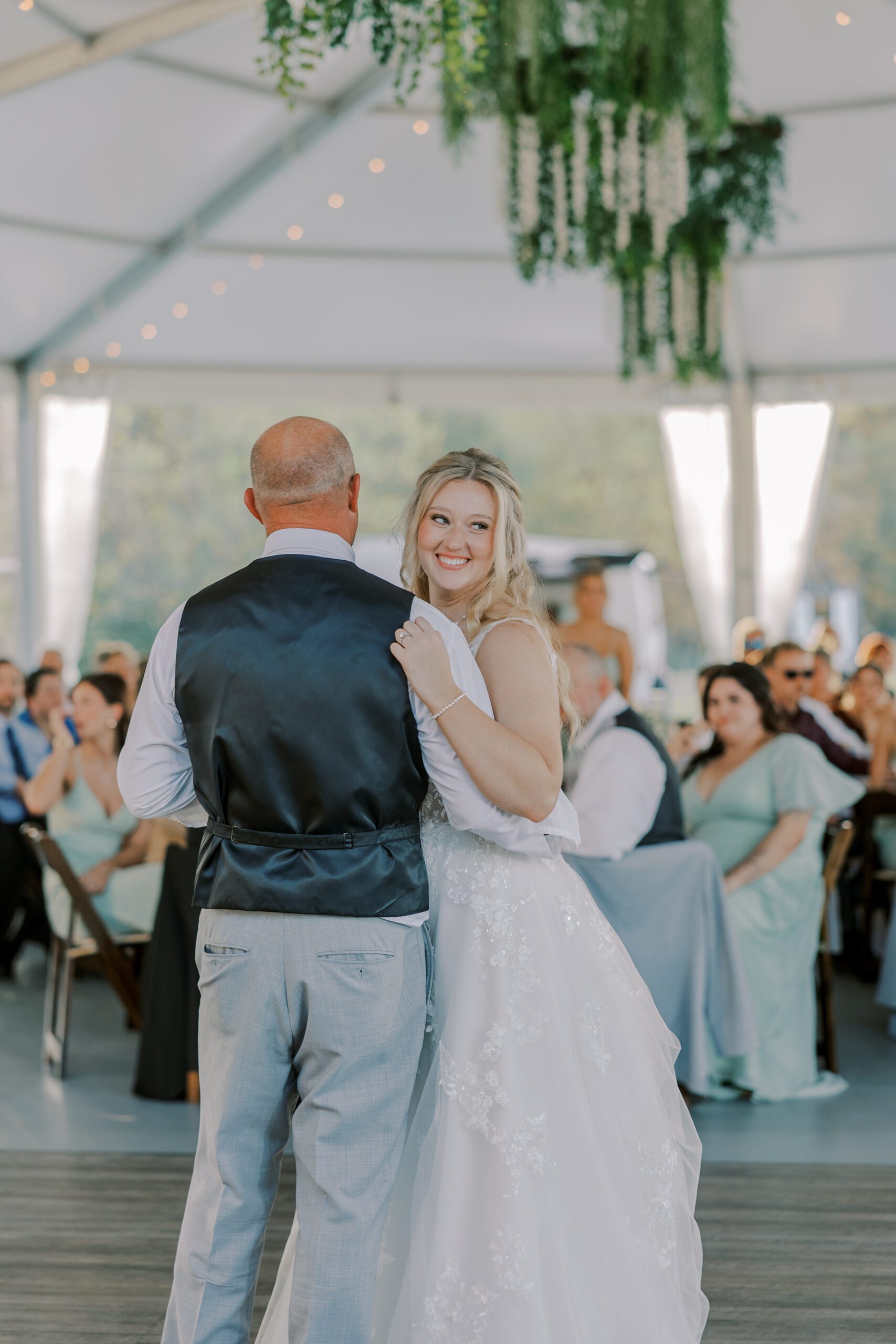 Bride dancing with her father, who is facing away from camera. Bride looks off to the side smiling, guests behind them sitting at tables