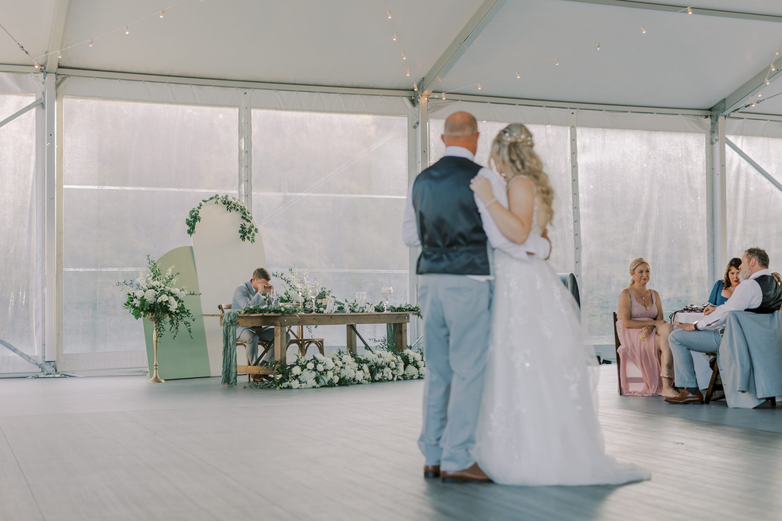 Bride and her father, out of focus dancing looking over at the groom who is in focus, clearly emotional with his face in his hands