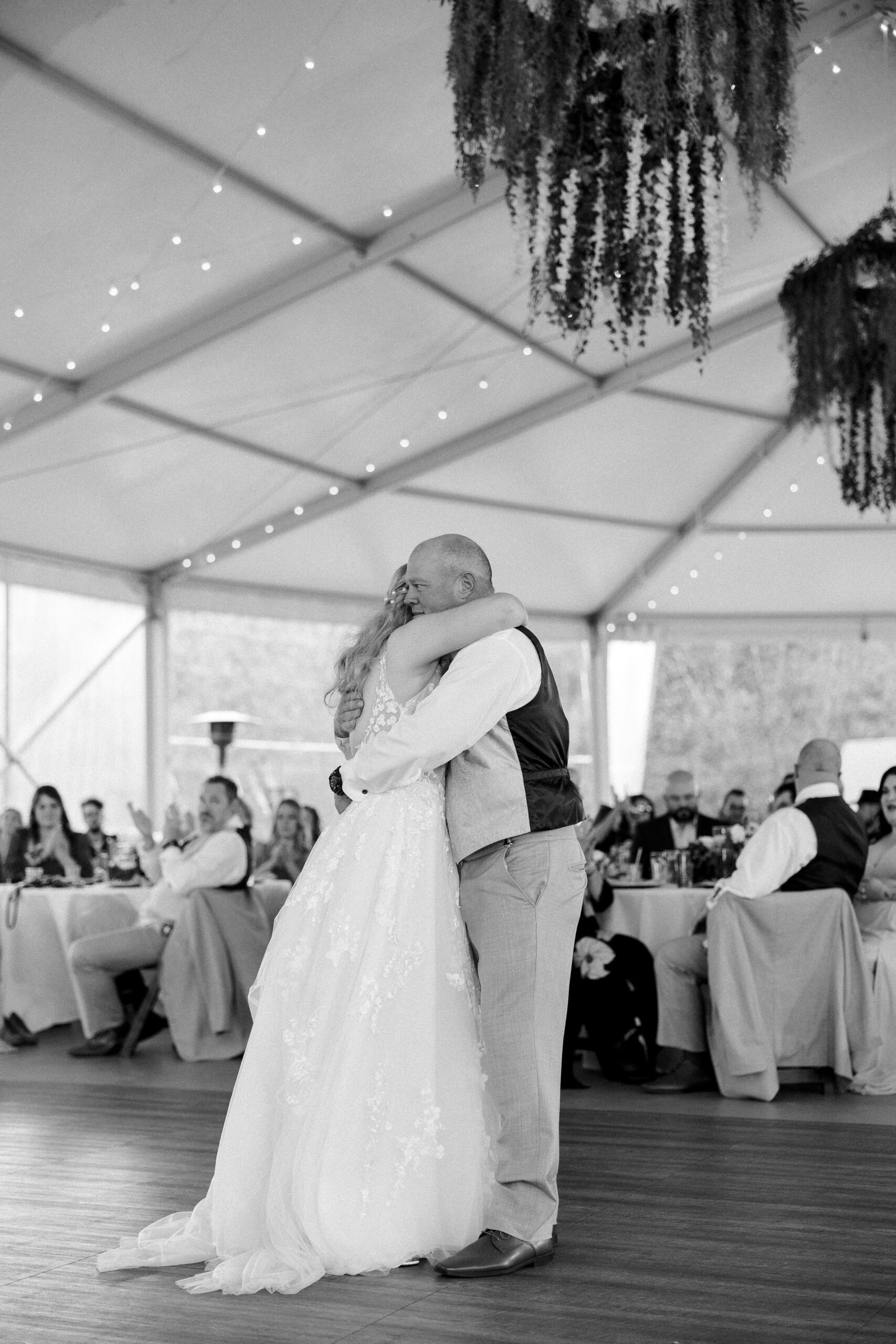 Black and white photo of bride hugging her father on the dance floor , twinkle lights and greenery hang above