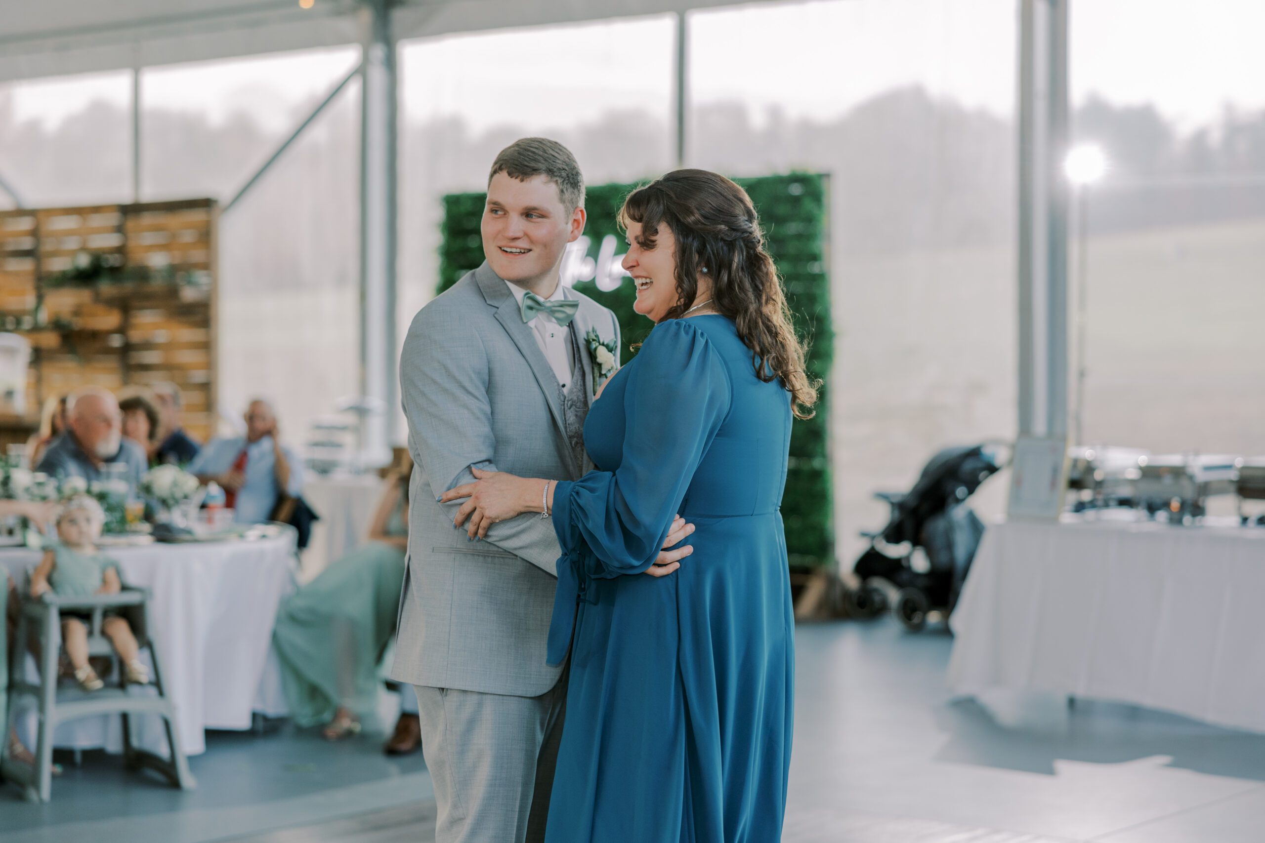 Groom and his mother dancing, both looking off to the side smiling, mother is in long sleeve blue dress