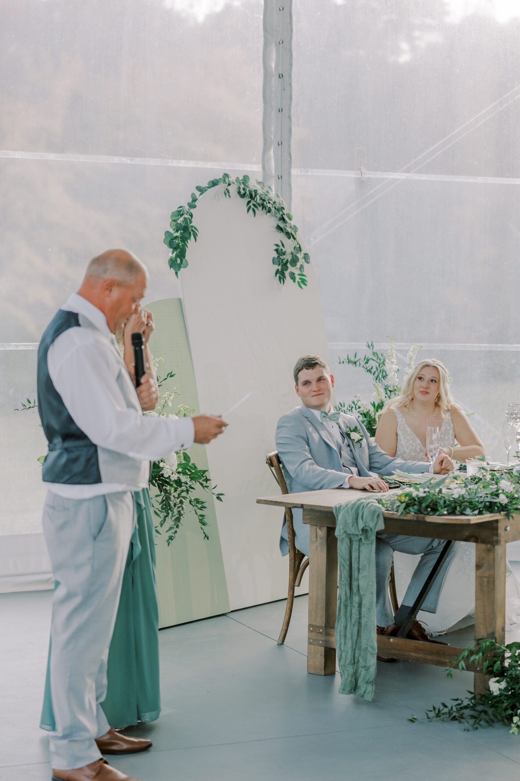 Father of the bride giving a speech while bride and groom watch from their table, white and green decorative arched pieces behind them with greenery on them