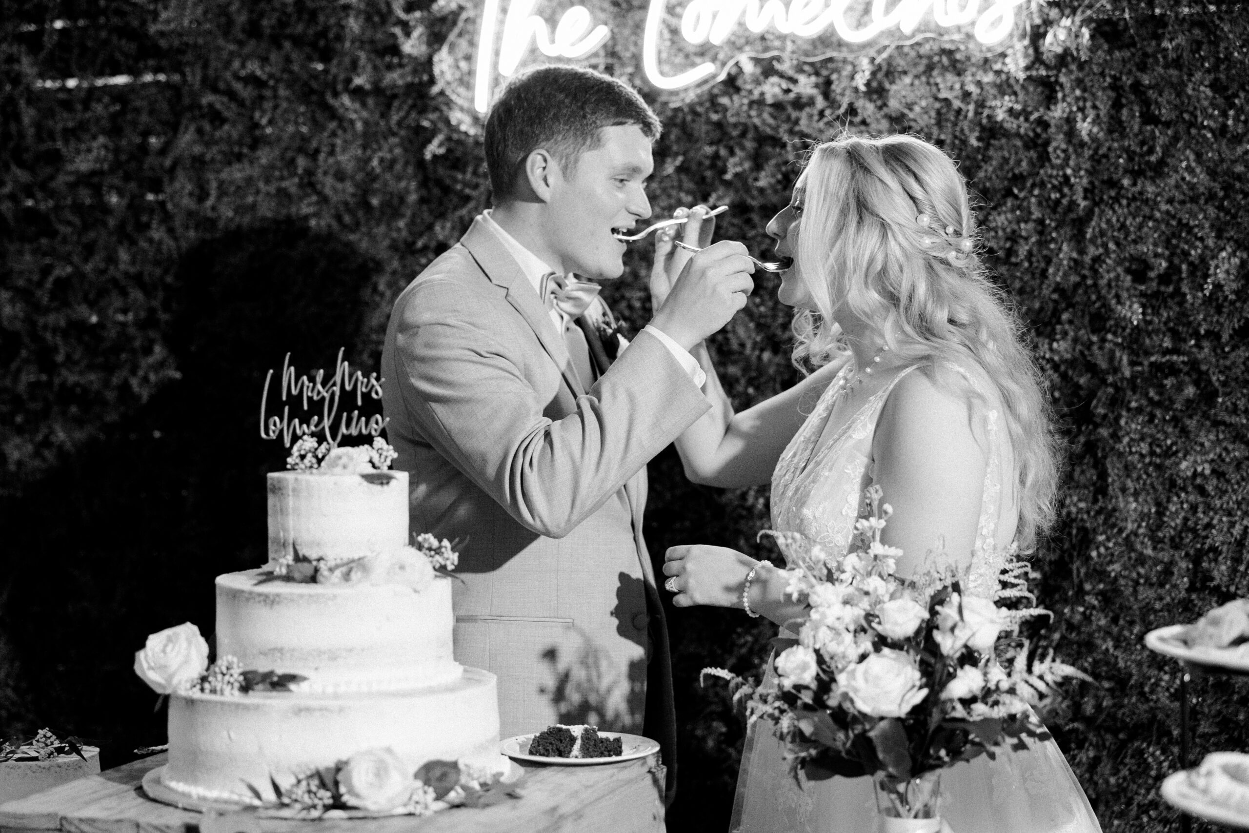 Black and white photo of bride and groom standing next to their cake, feeding each other a bite with forks, light up name sign in background