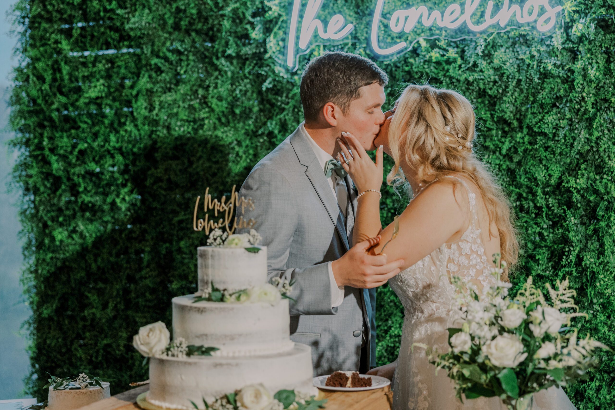 Bride and groom kissing next to their wedding cake at their arbor haven fall wedding, greenery wall with light up last name sign pictured behind them