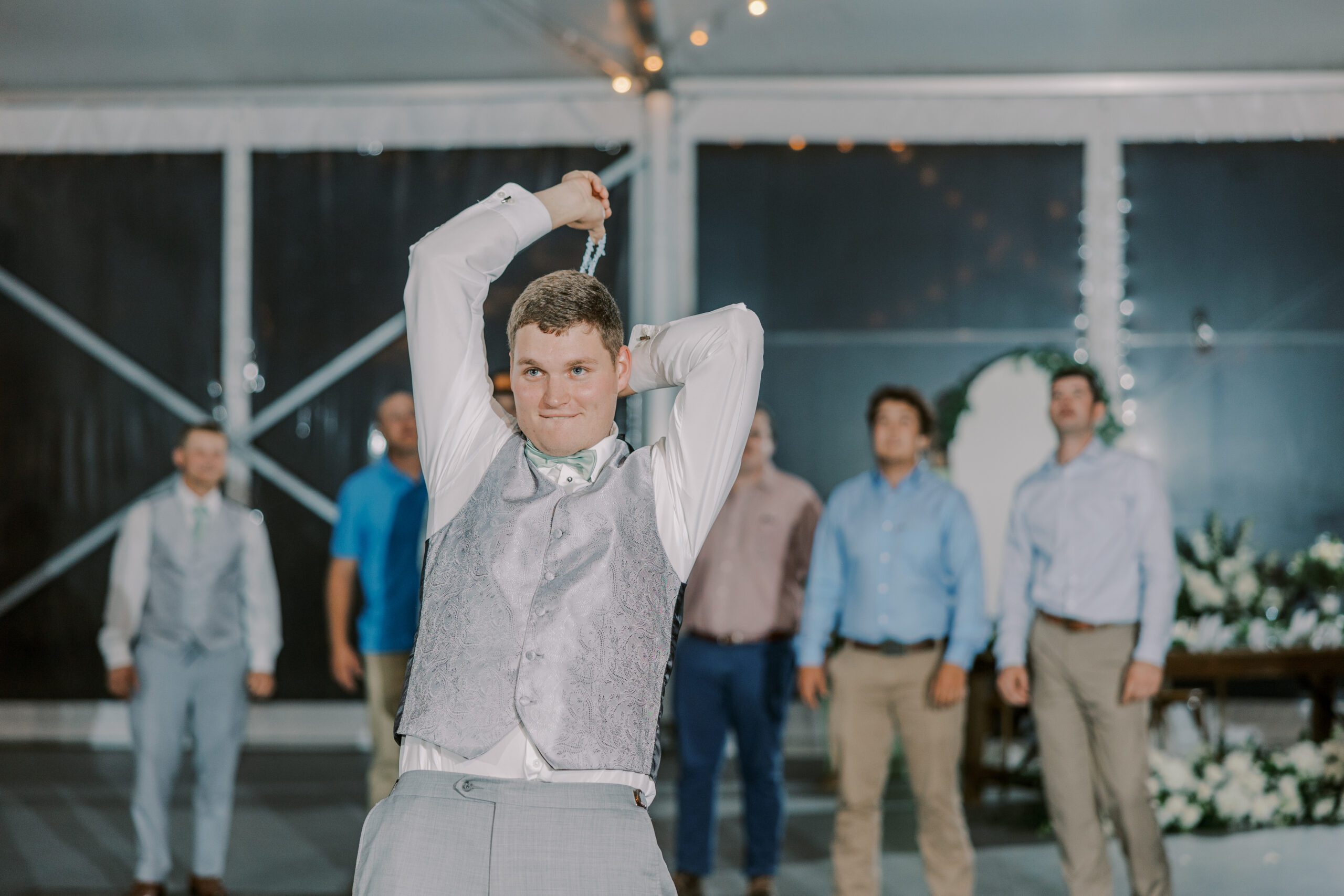 Groom in focus getting ready to toss the garter behind him, as groomsmen and male guests can be seen standing behind him