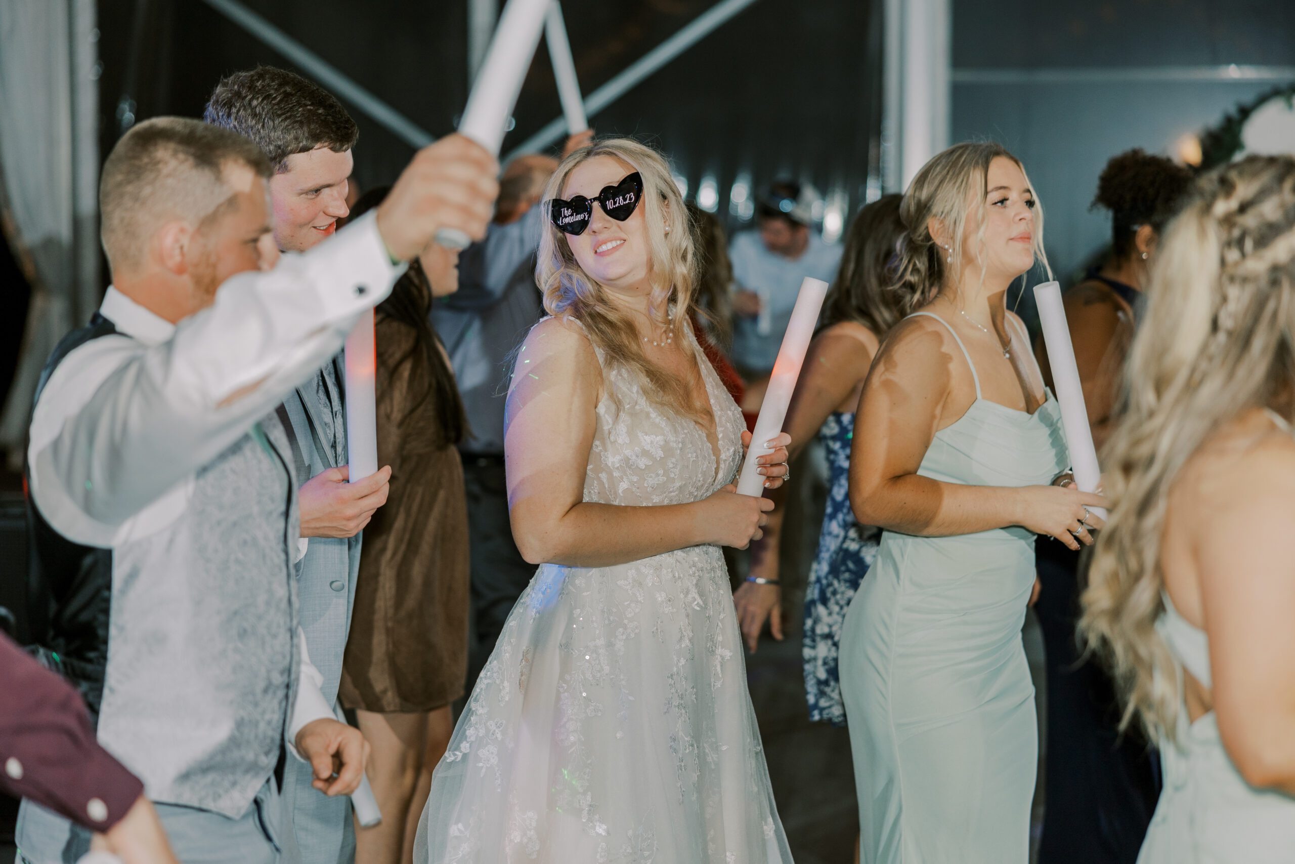 Bride wearing heart shaped sunglasses with her new last name and wedding date written on them, holding a light up baton and dancing alongside other guests