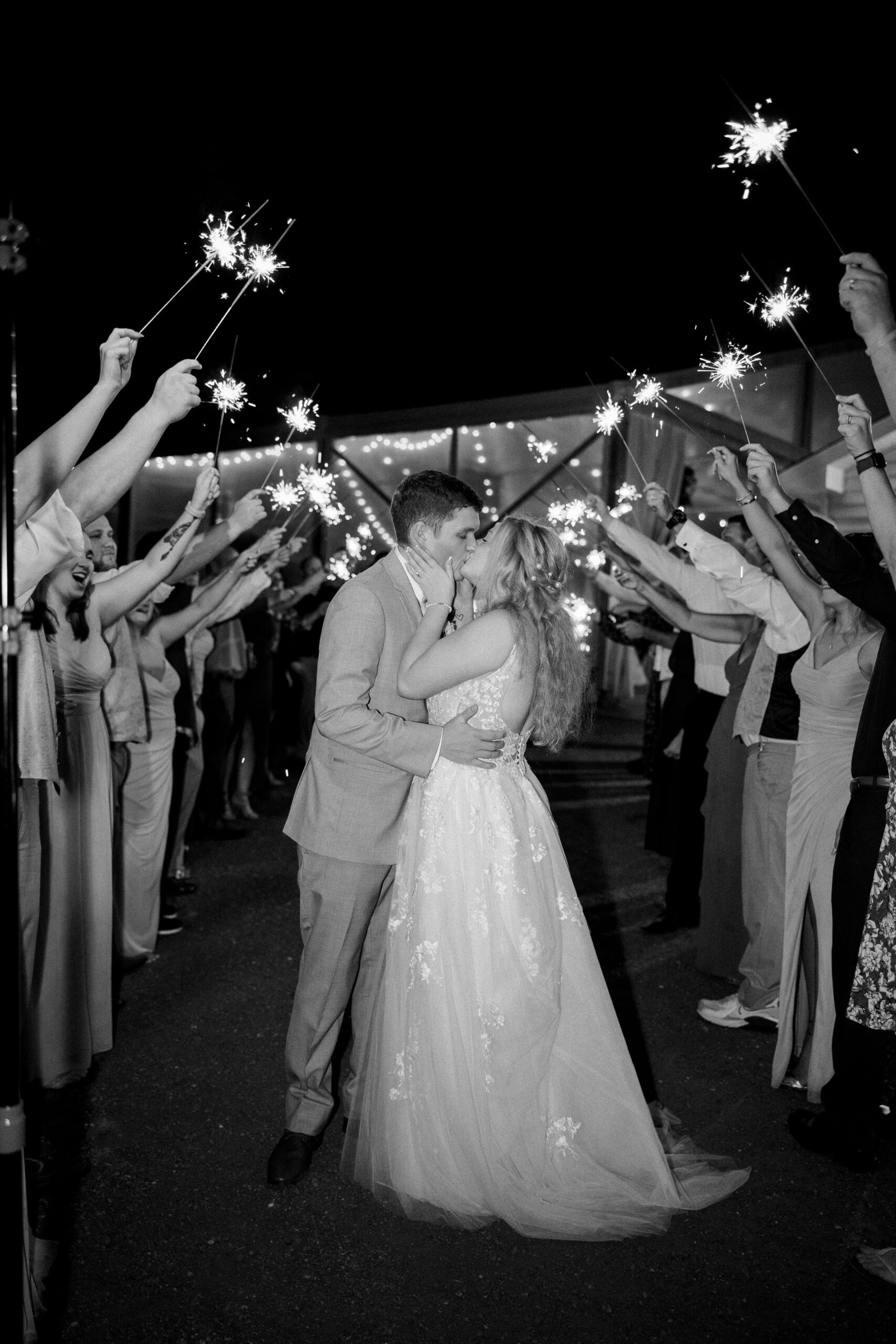 Black and white image of bride and groom kissing as guest hold up sparklers around them at their arbor haven fall wedding