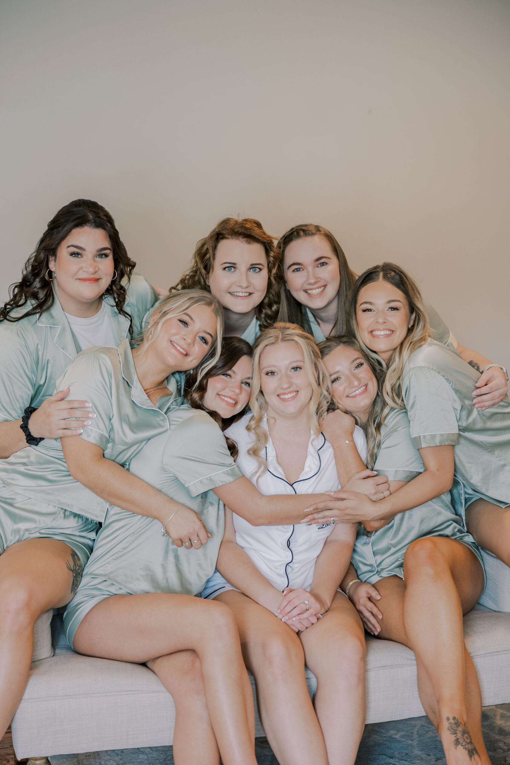 Bride in white pajamas with 7 bridesmaids all in matching light green pajamas lean in hugging one another while they sit on a couch at arbor haven