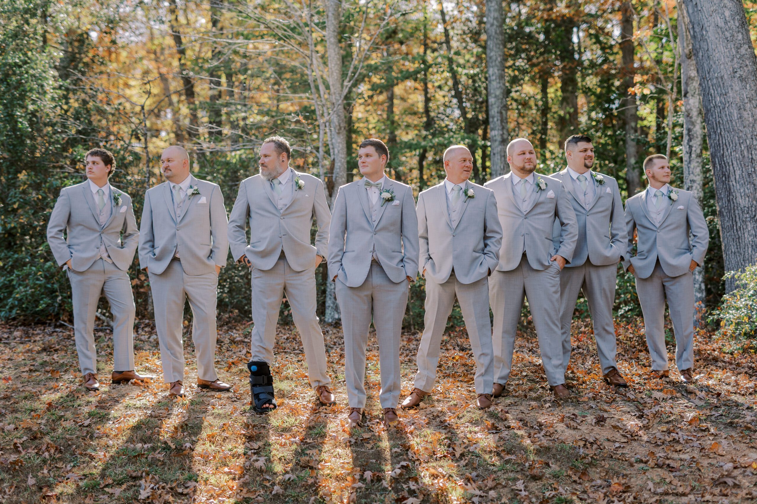 Groom and his 7 groomsmen standing in a wooded area, trees behind them and fall leaves on ground. they are all wearing light grey suits