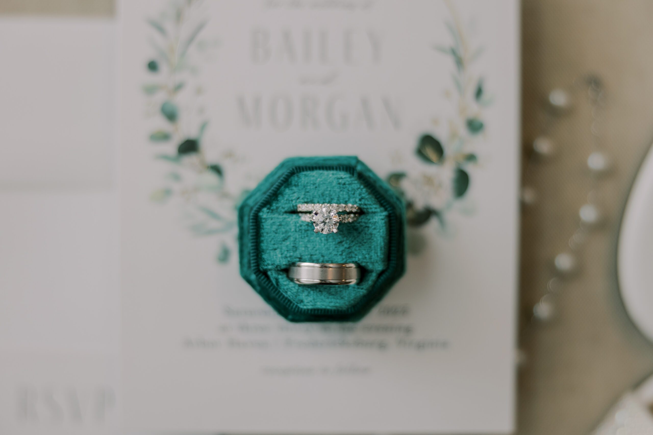 Close up photo of bride and groom's rings in an octagon shaped green velvet ring box, invitation is out of focus in background