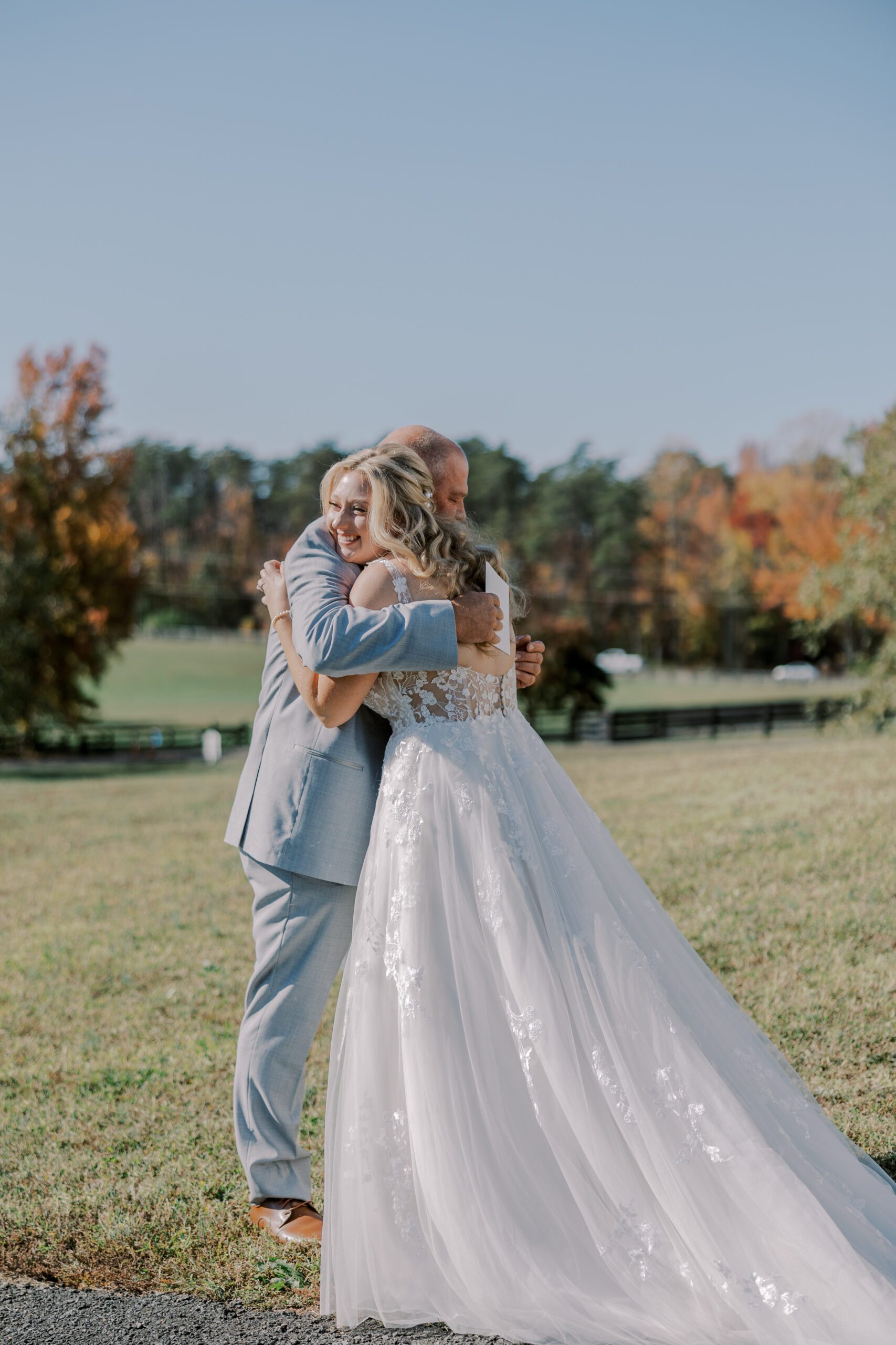 Full length image of bride and her father hugging, back of bride's dress pictured with lots of lace detailing scattered down the skirt of the dress