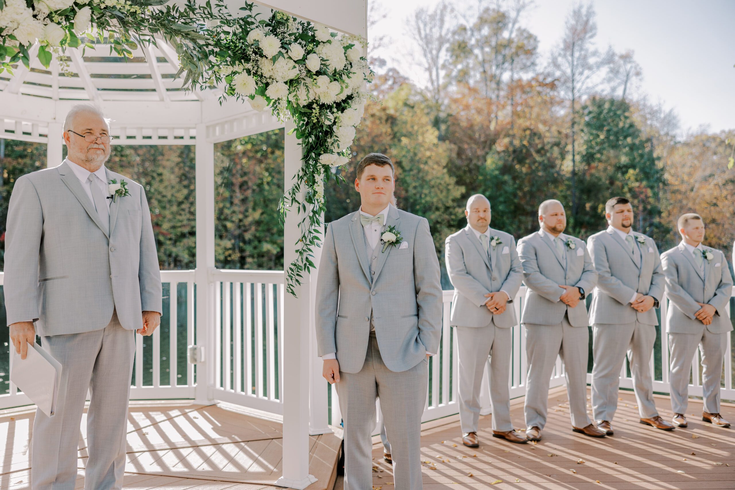 Groom standing next to the gazebo, officiant and groomsmen on either side of him, there are white florals hanging from gazebo at the arbor haven fall wedding