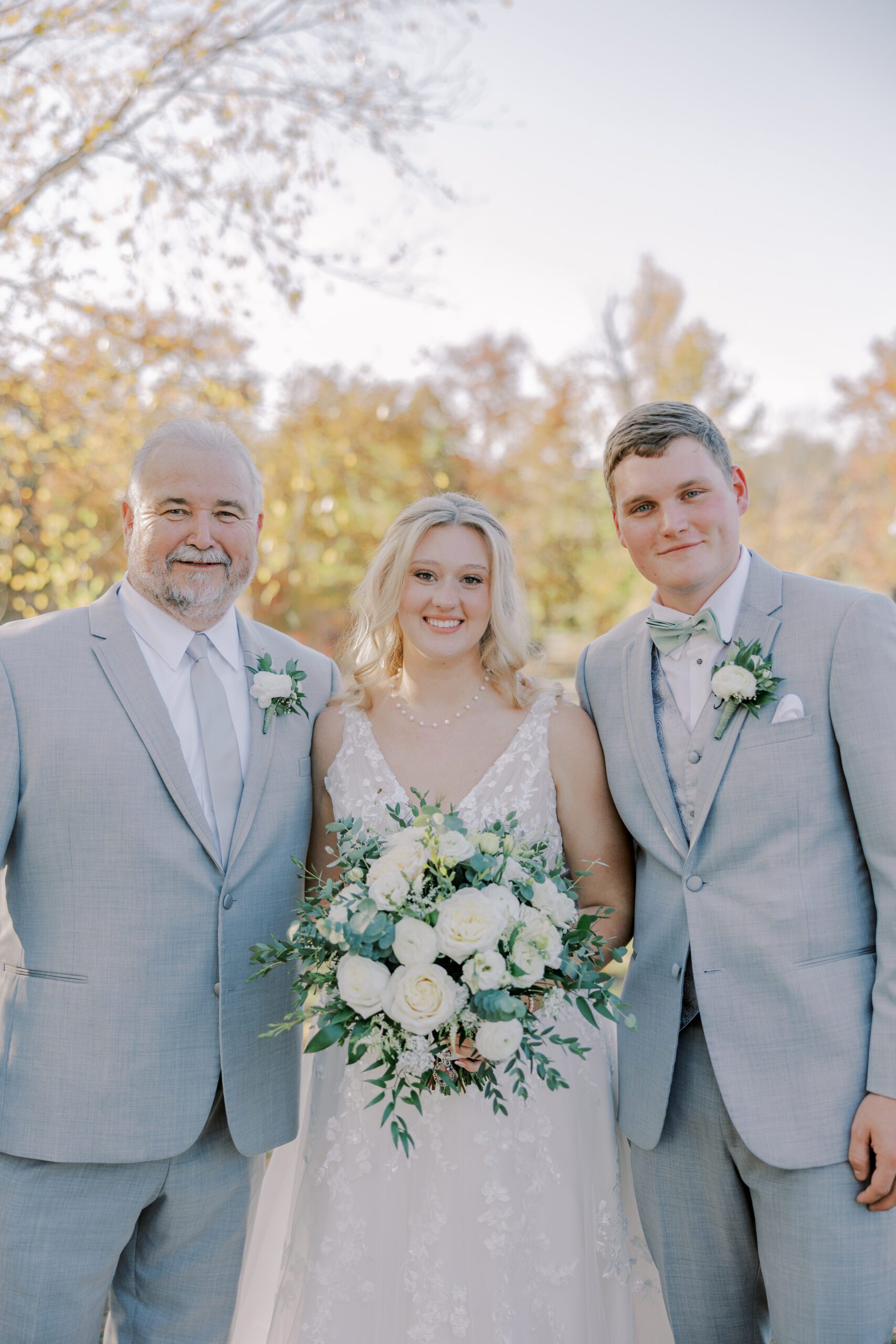 Bride and groom pictured with their officiant, bride in middle holding her bouquet of flowers, all 3 people looking and smiling at camera