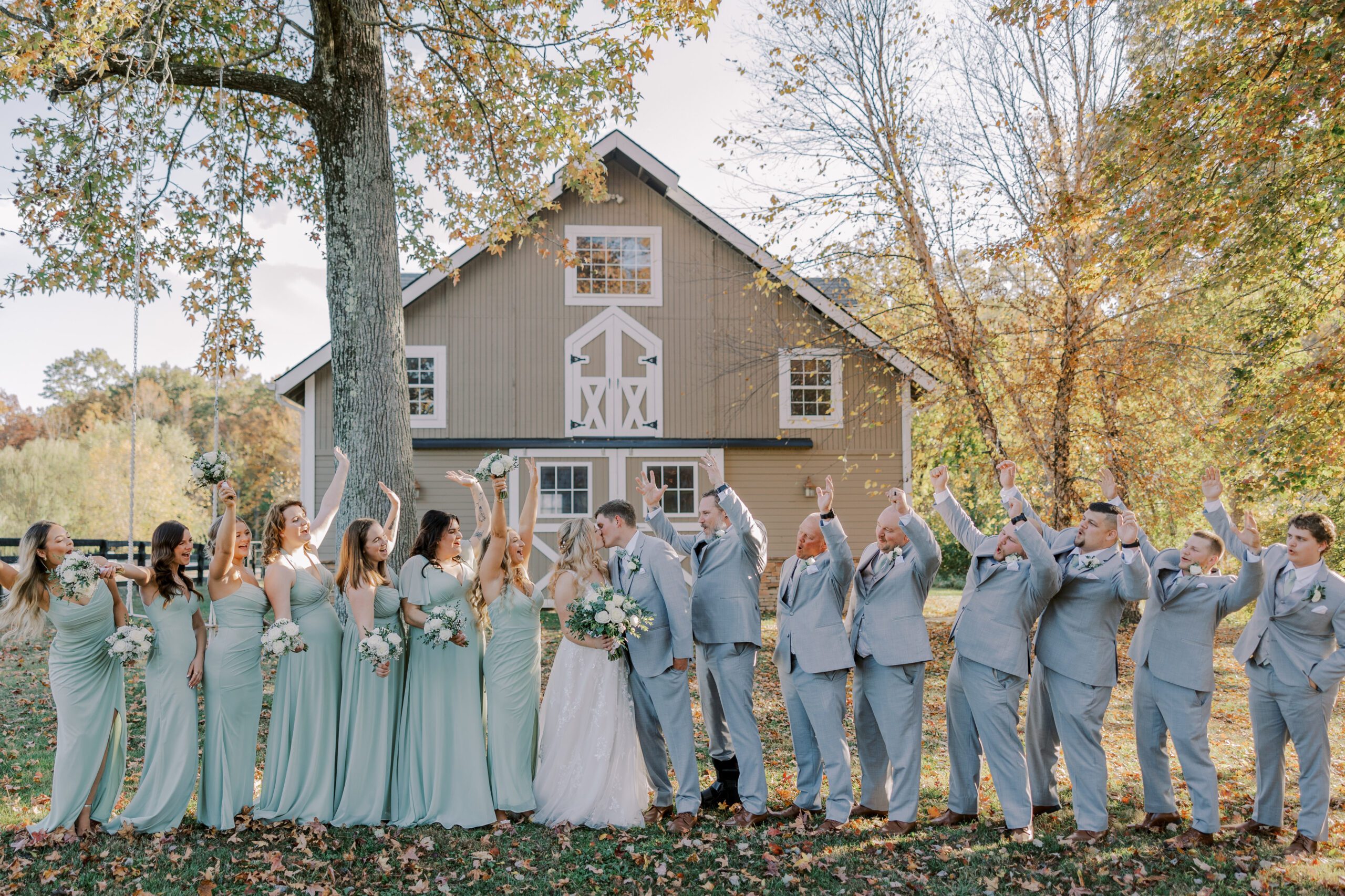 Wedding party standing in line, bride and groom in center kissing while everyone else cheers them on, light brown barn and trees are in background of photo