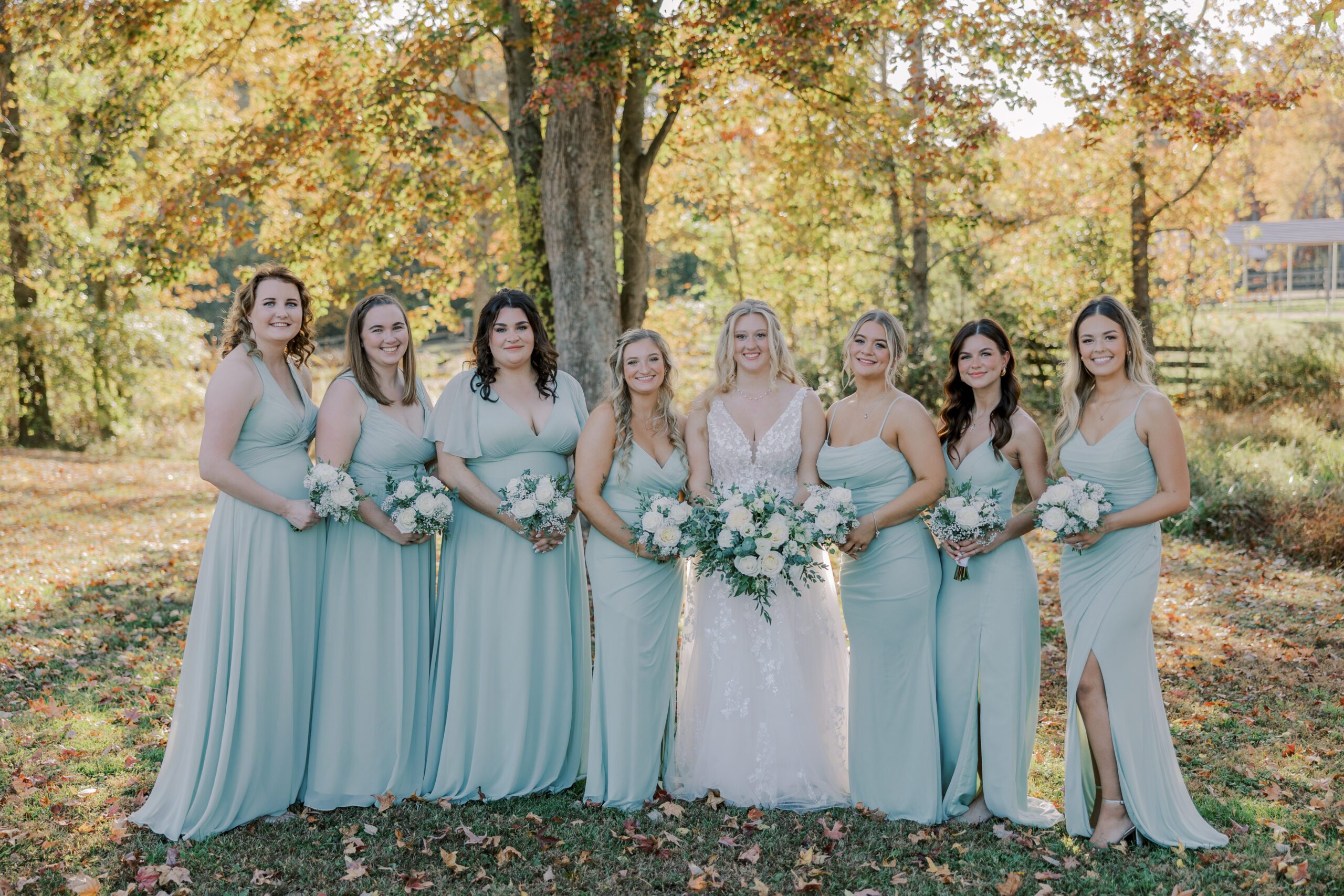 Bride and her 7 bridesmaids all holding bouquets of white flowers, wearing light green dresses, smiling, fall colored trees in background at arbor haven