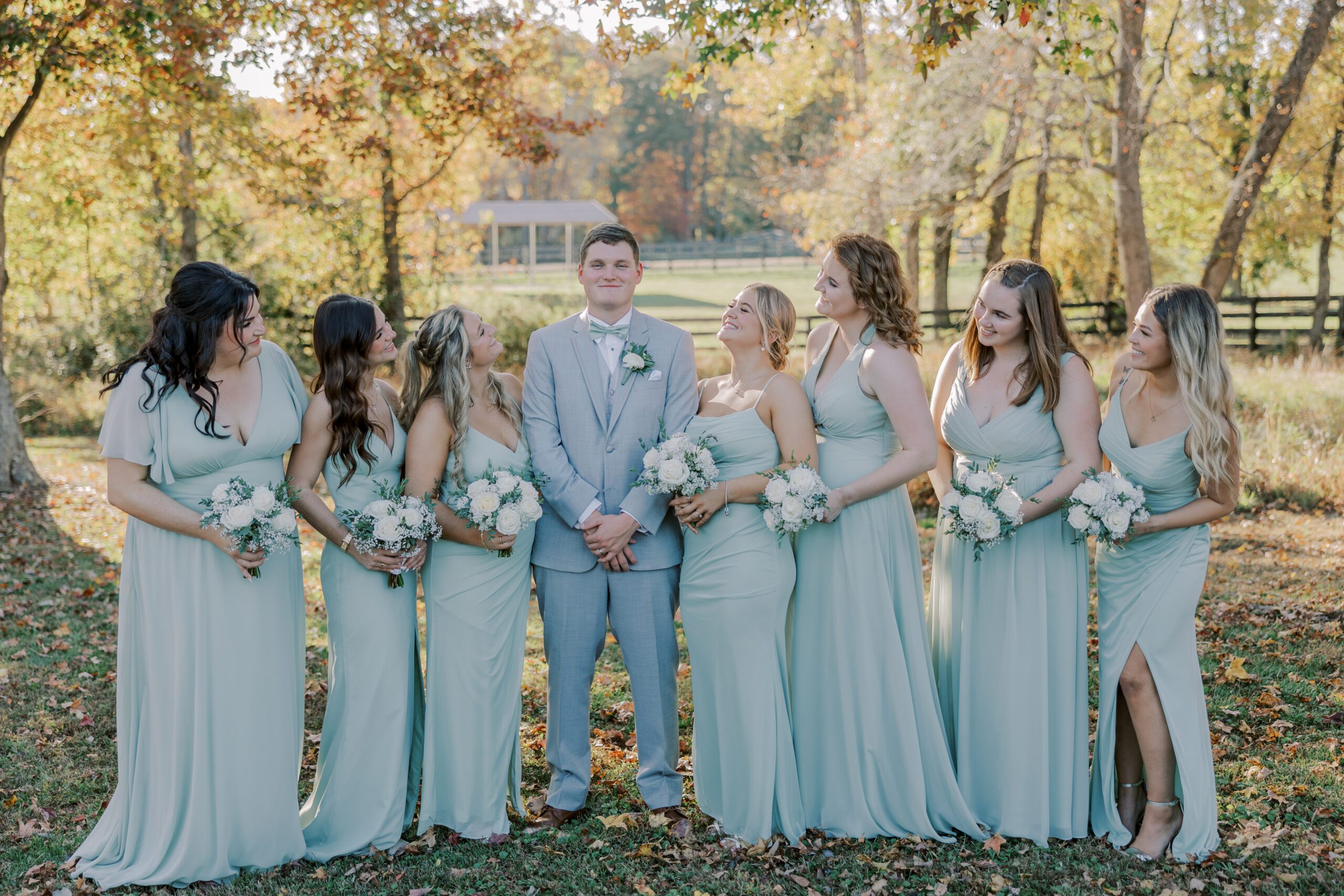 Groom standing in middle of line of bridesmaids who are all smiling at him while he looks forward also smiling