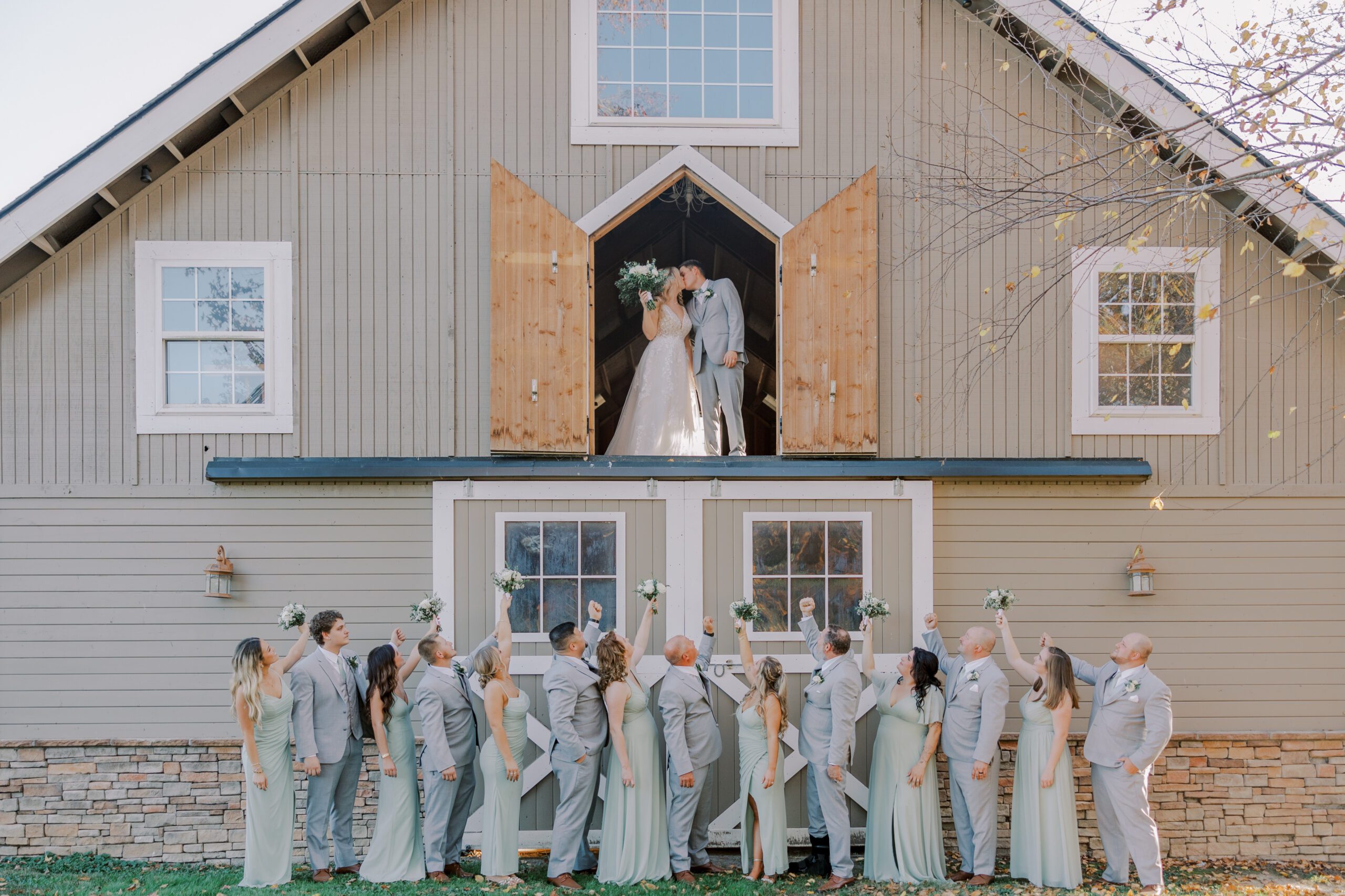 Bride and groom stand and share a kiss on the second floor of light brown barn while their wedding party is outside looking up at them, arms raised at their arbor haven fall wedding