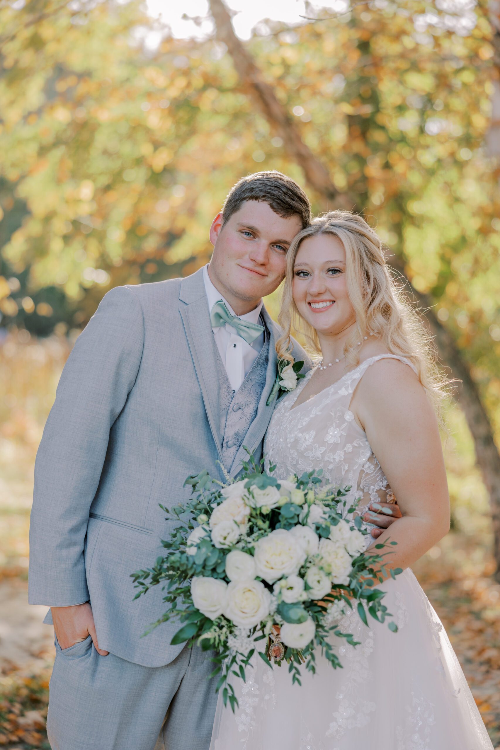 Image of bride and groom, autumn colored trees in background, both smiling at camera at their arbor haven fall wedding