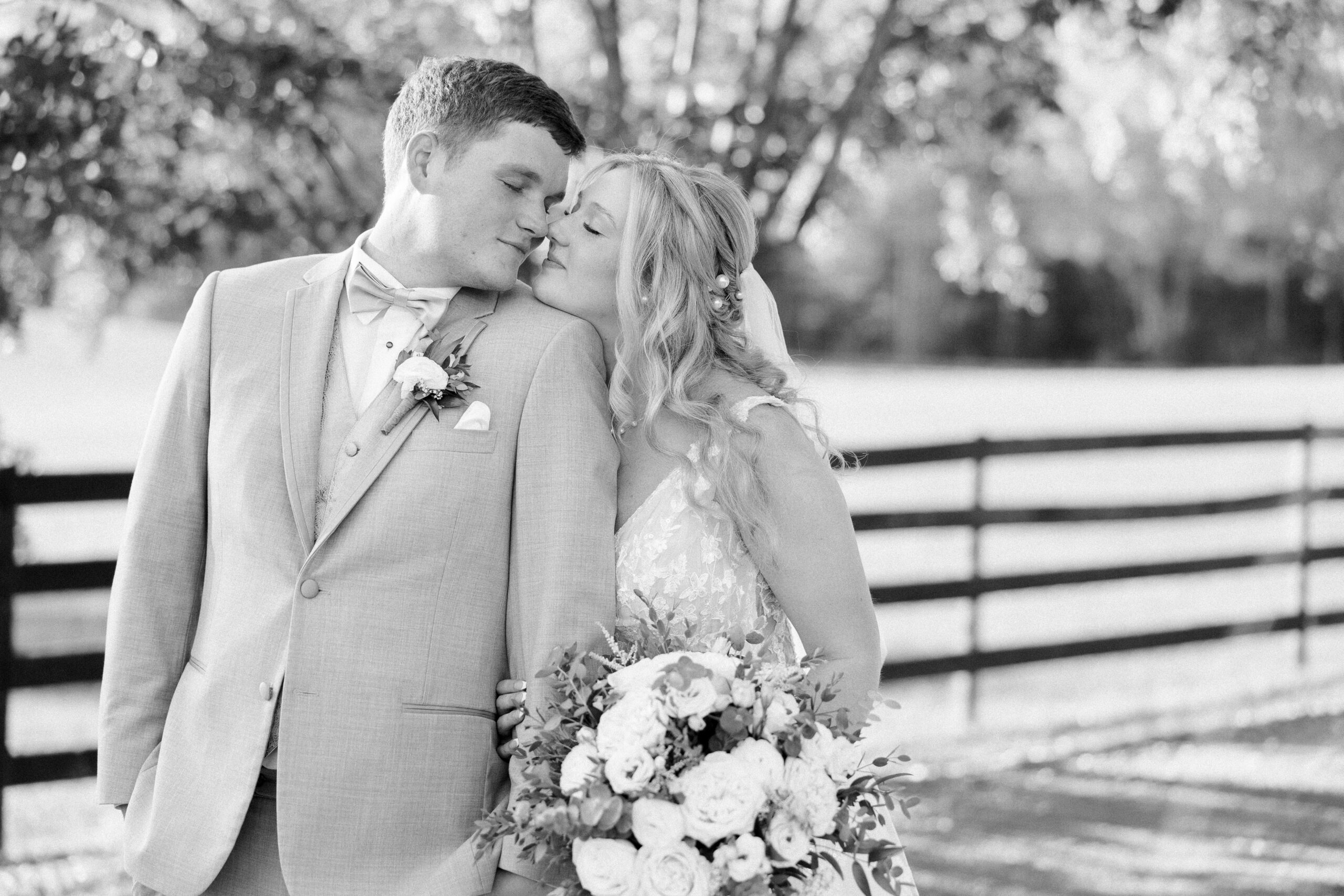 Black and white photo of bride standing behind groom with her chin on his shoulder, they're touching noses with their eyes closed, fence and tress can be seen behind them at arbor haven