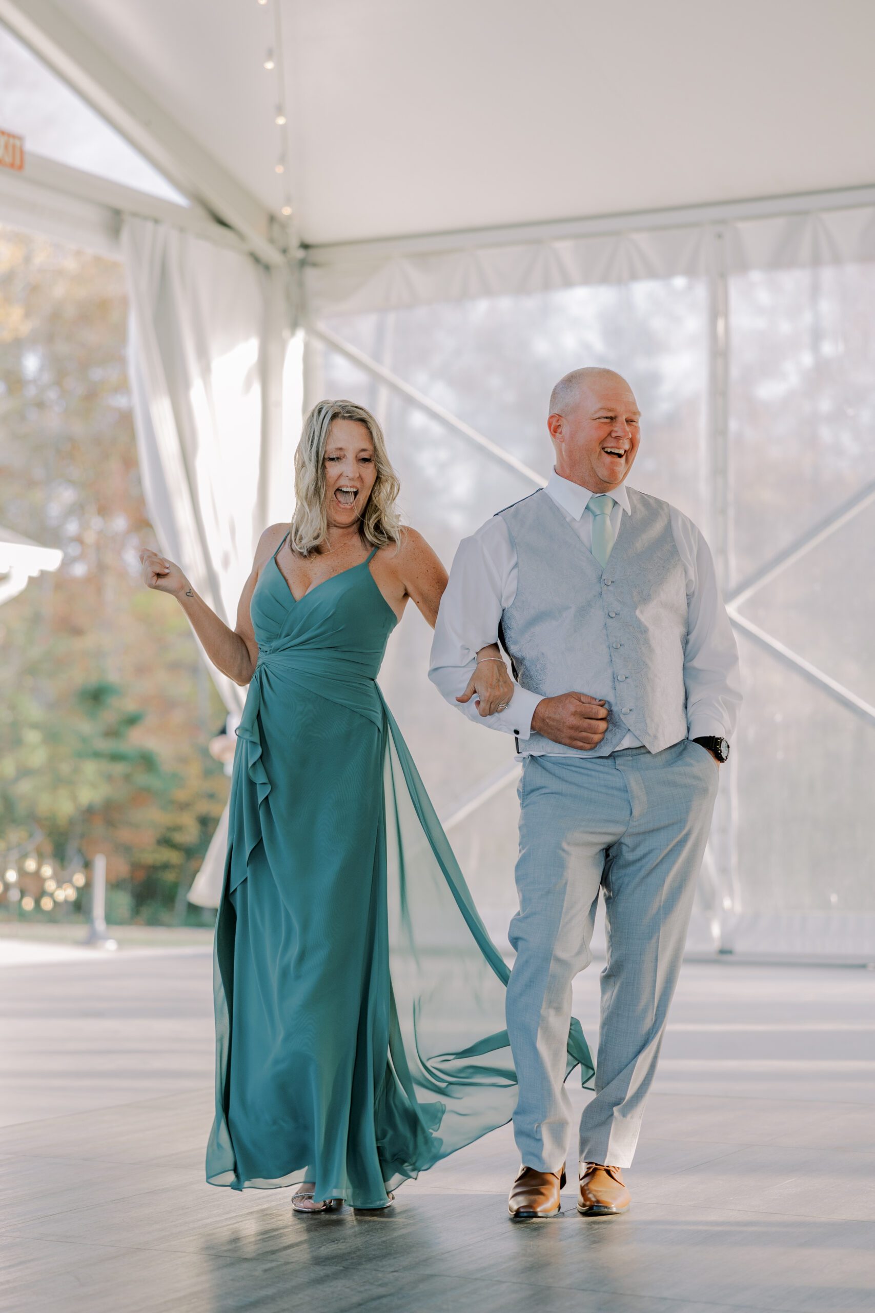 Bride's parents enter reception smiling and laughing, woman is wearing a dark teal colored dress and man is in light grey suit without the jacket