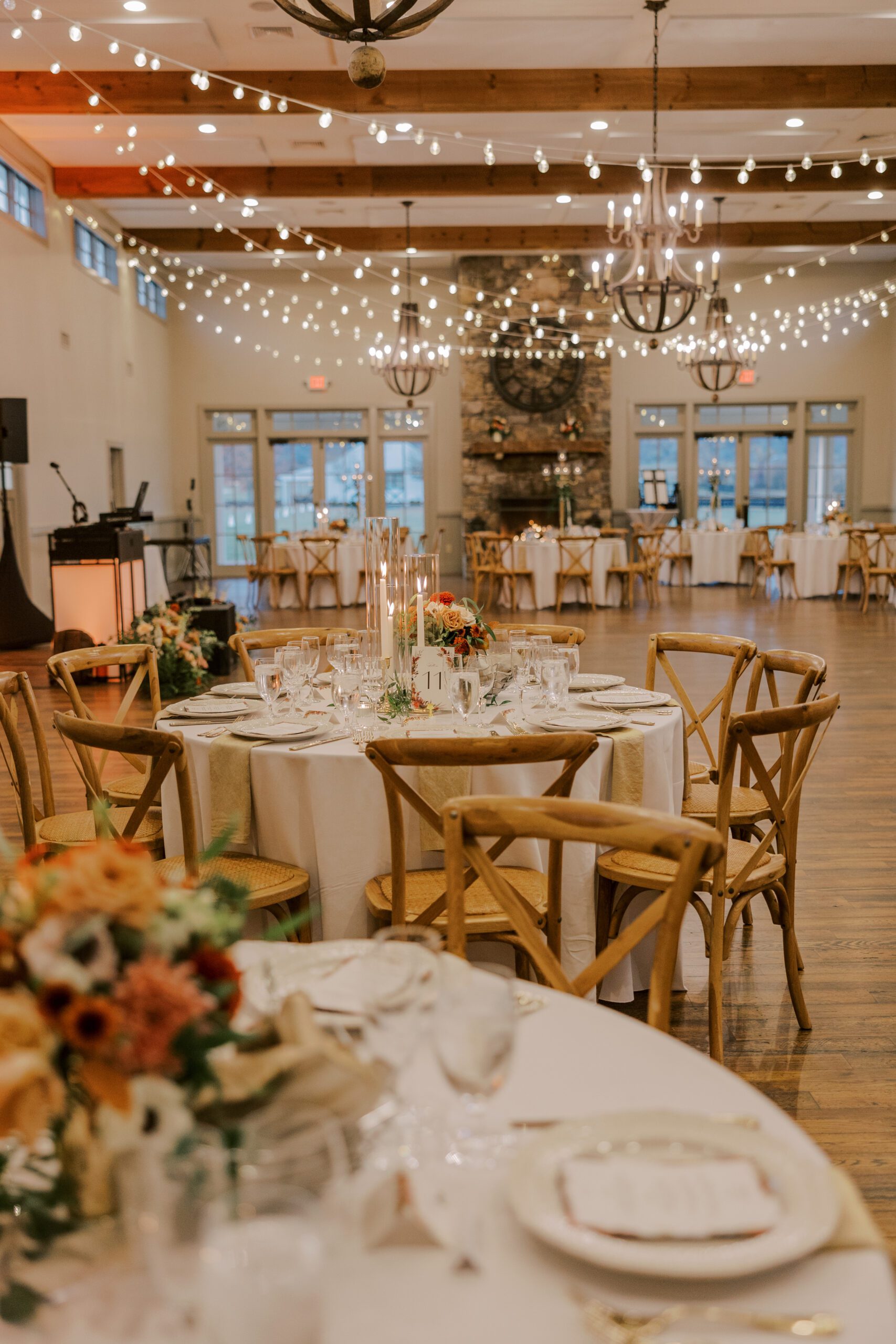 Tables at the reception with light wooden chairs, fall colored flowers and candles as the centerpieces, with string lighting overhead