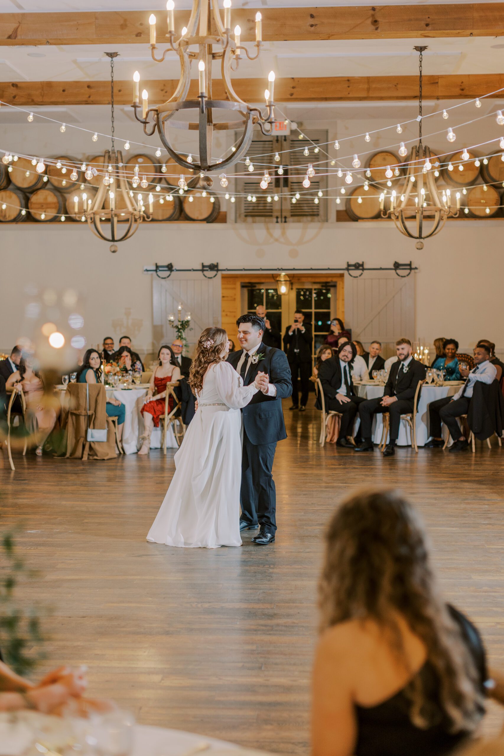Bride and groom share a dance in their reception as guests sit at tables watching, chandelier and string lights hand overhead