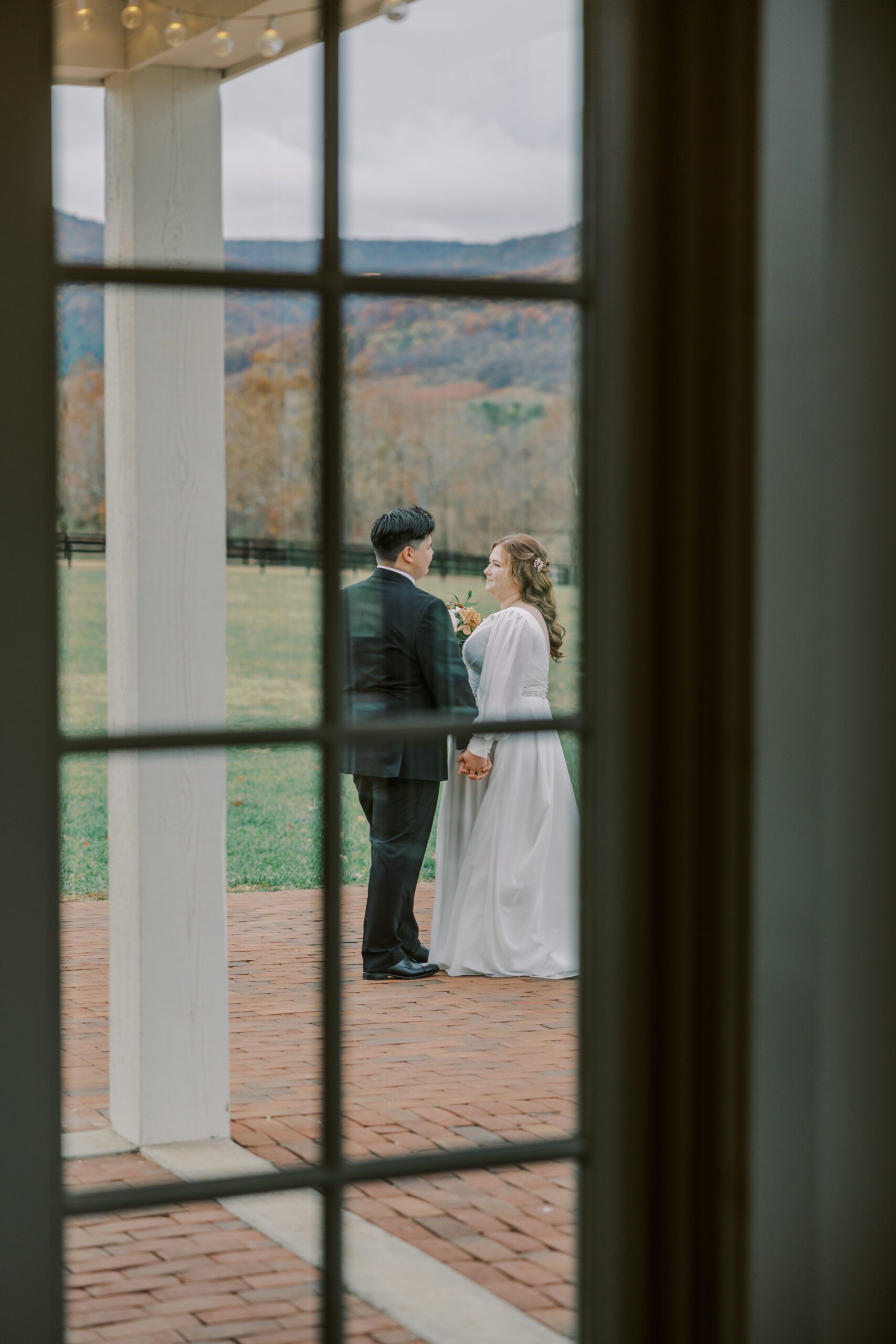 Distance photo taken of bride and groom through a window as they stand outside sharing an quite moment before their wedding ceremony