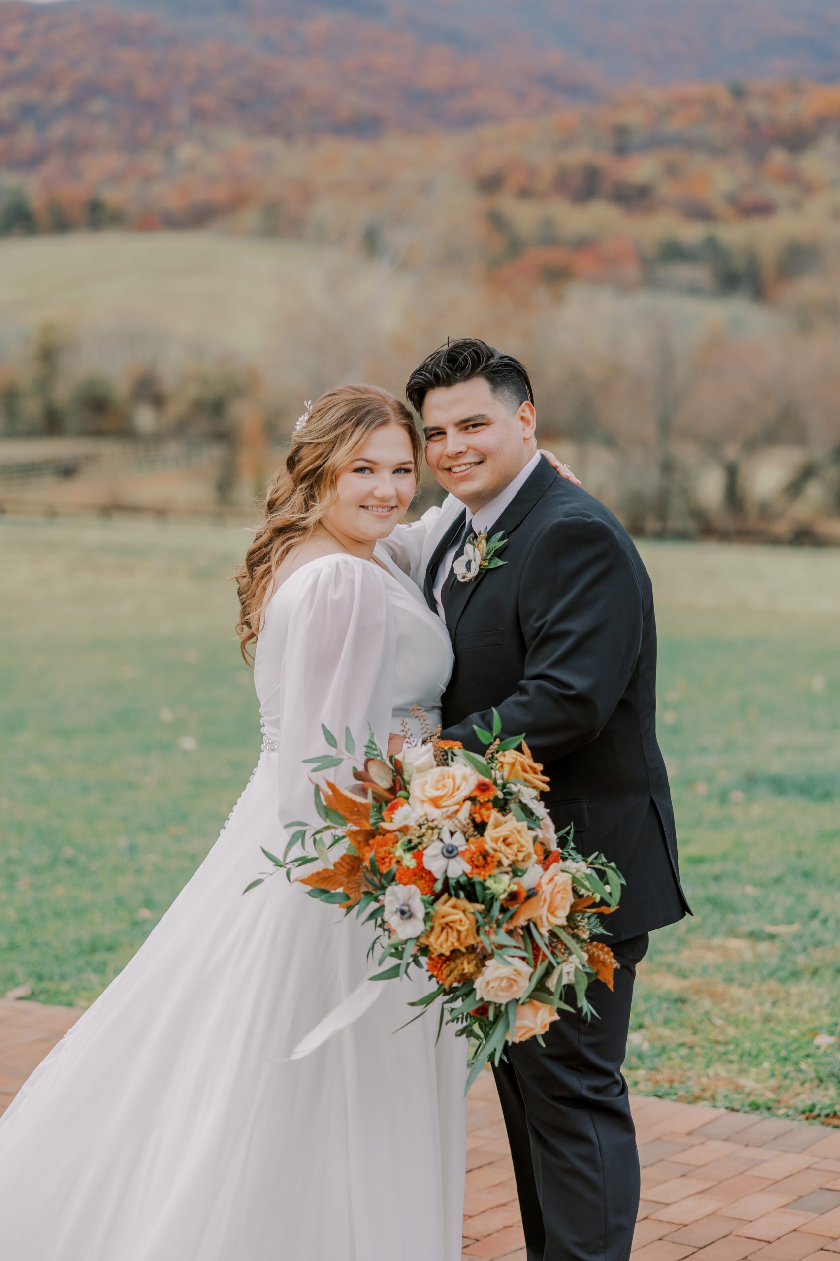 Bride and groom smiling at camera, standing outside with bride holding large floral bouquet with many white, peach, orange and green colors in it