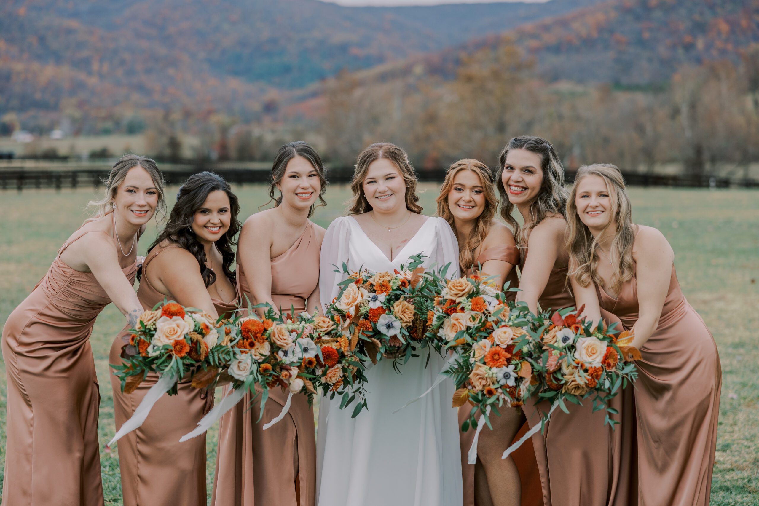 Outdoor photo of bride with her bridesmaids who are wearing light brown dresses, all holding their bouquets together and smiling at camera