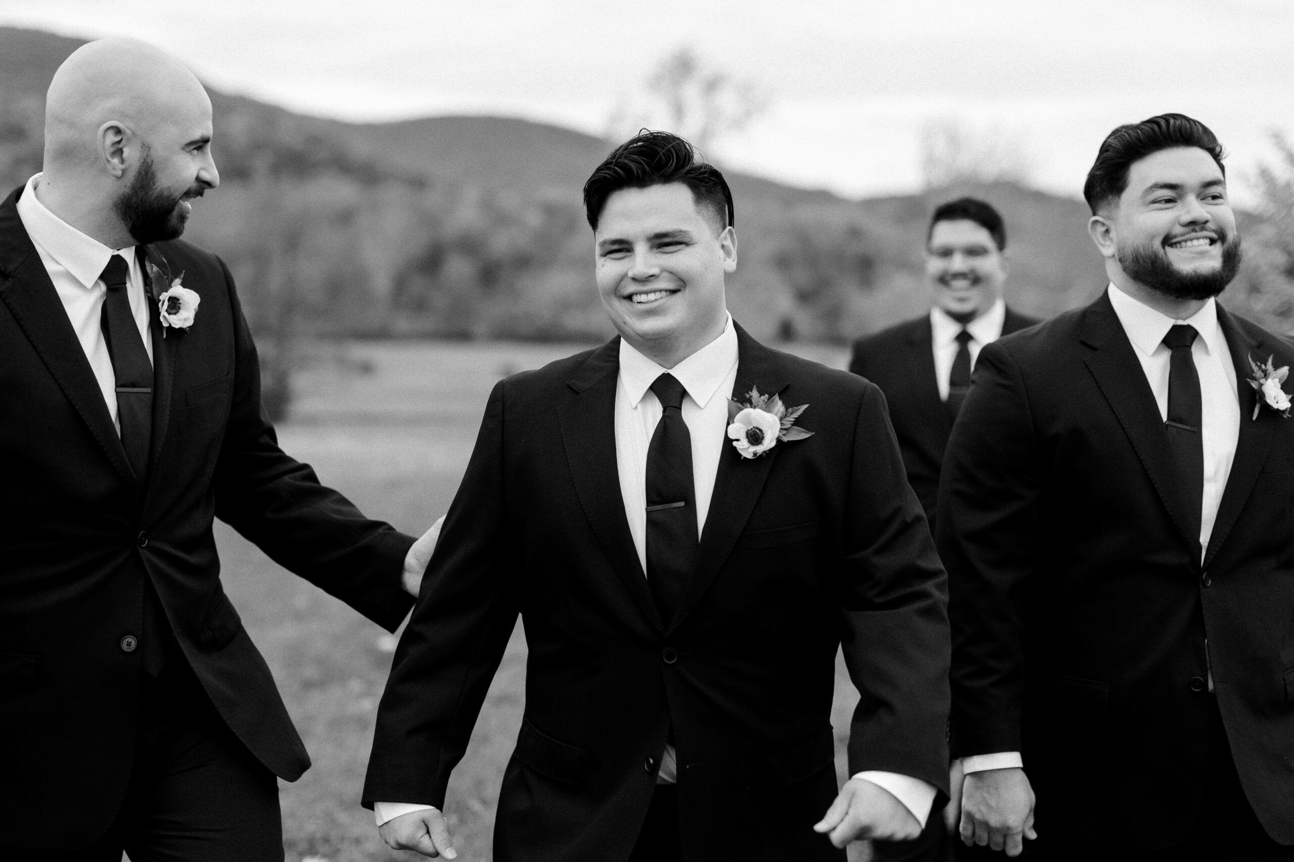Black and white image of groom walking, smiling, and a couple of his groomsmen are in the background