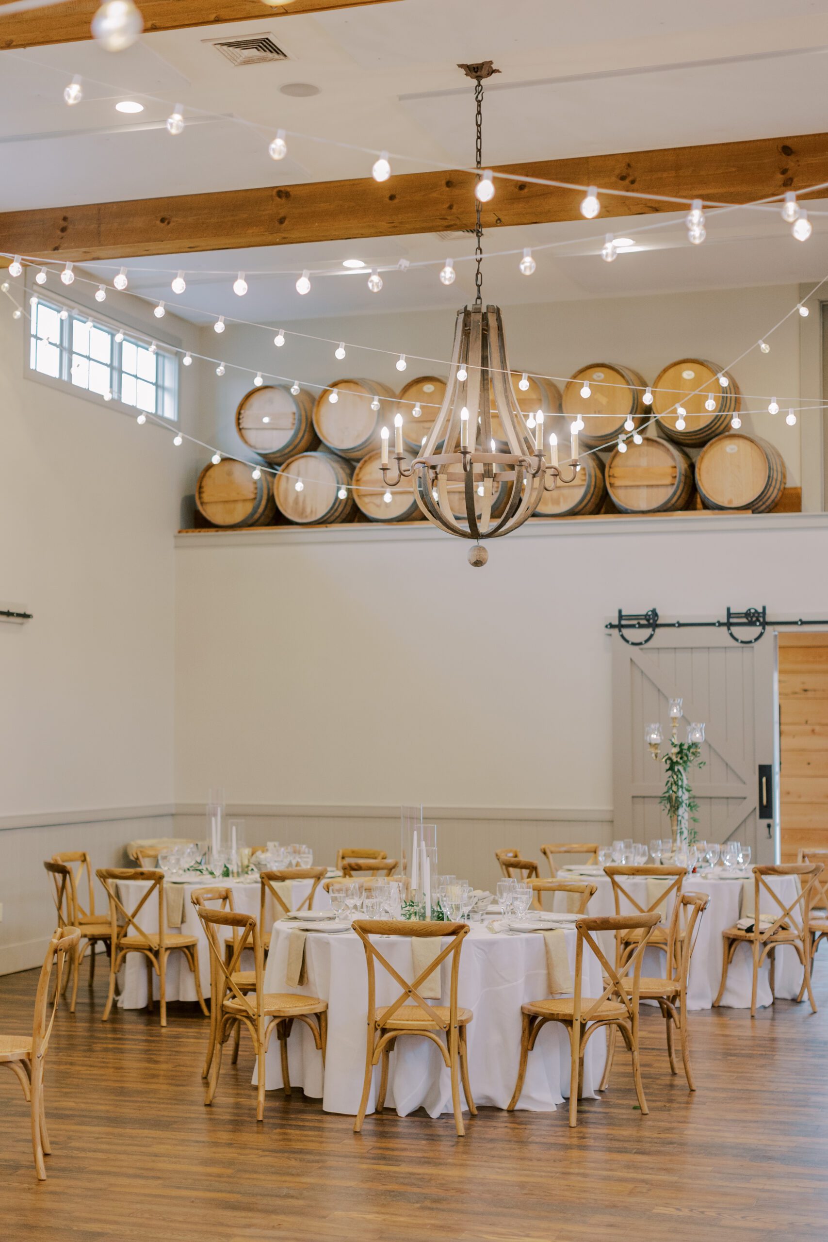 Image of reception space at king family vineyards with white table cloth covered tables, light wooden chairs, twinkle lights and barrels up on a high shelf