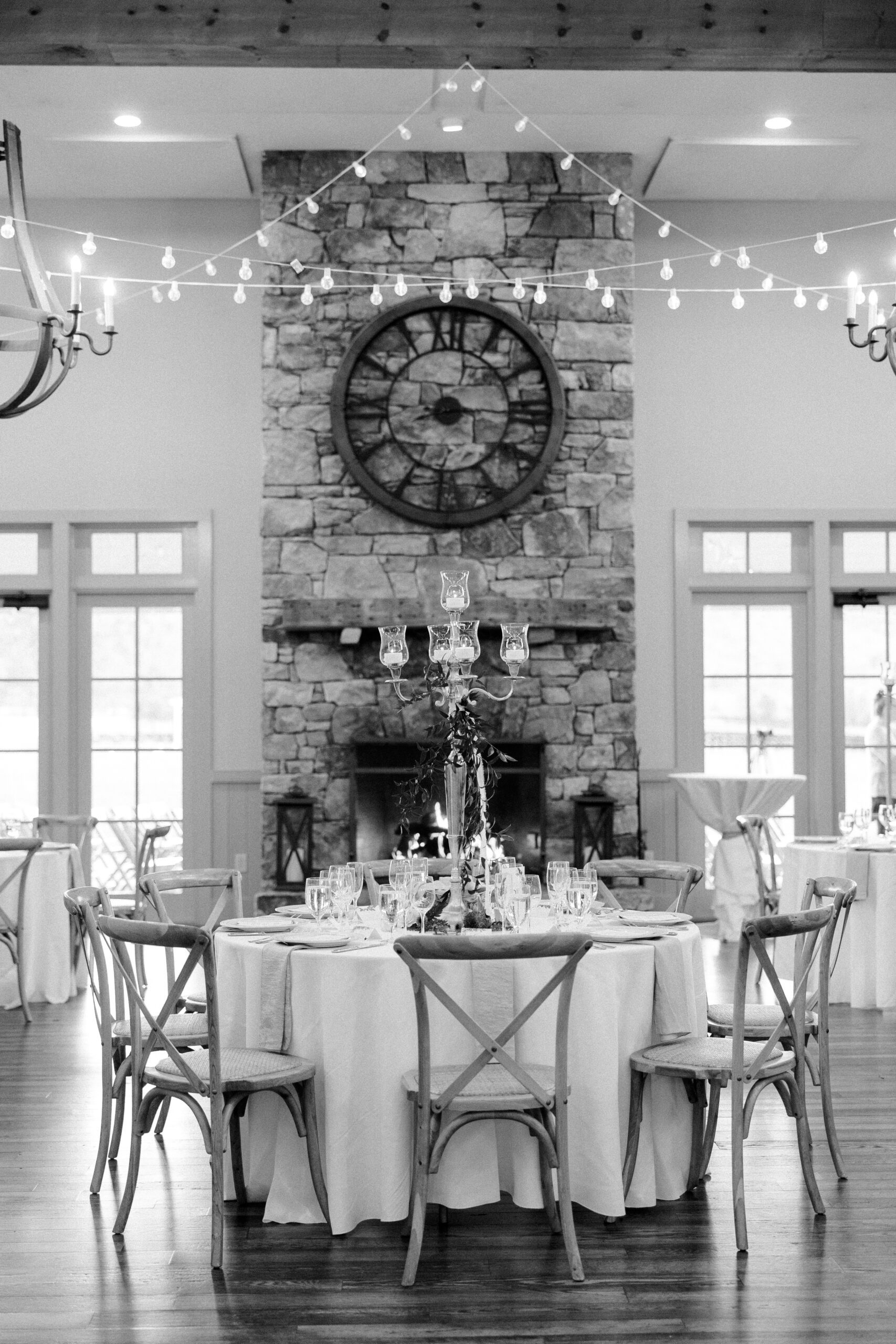 Black and white photo of reception space at king family vineyards showing a table, twinkle lights, and a clock on a stone fireplace wall